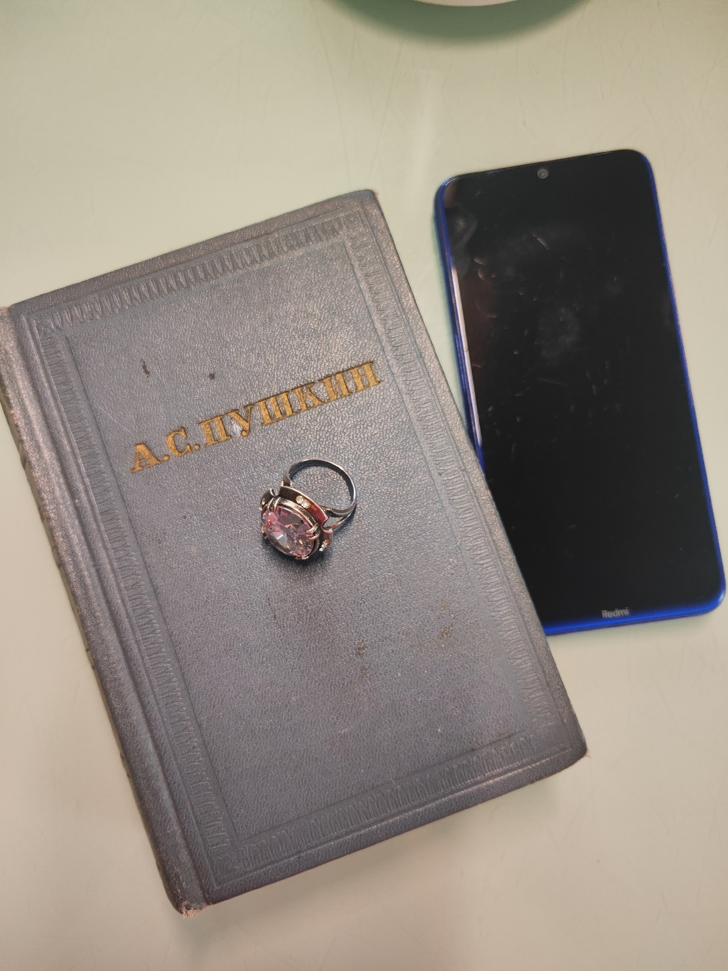 The image shows a book with gold lettering that reads "А. С. ПУШКИН" (A. S. Pushkin) lying on a light-colored surface. Placed on top of the book is a ring with a large pink gemstone and smaller clear stones. To the right of the book is a black smartphone with a blue border. The background is a light, textured surface. The scene appears to be an indoor setting, possibly a desk or table. No specific location within Gvardeysk, Russia can be determined from the visual information provided. There are no people or discernible actions in the image.