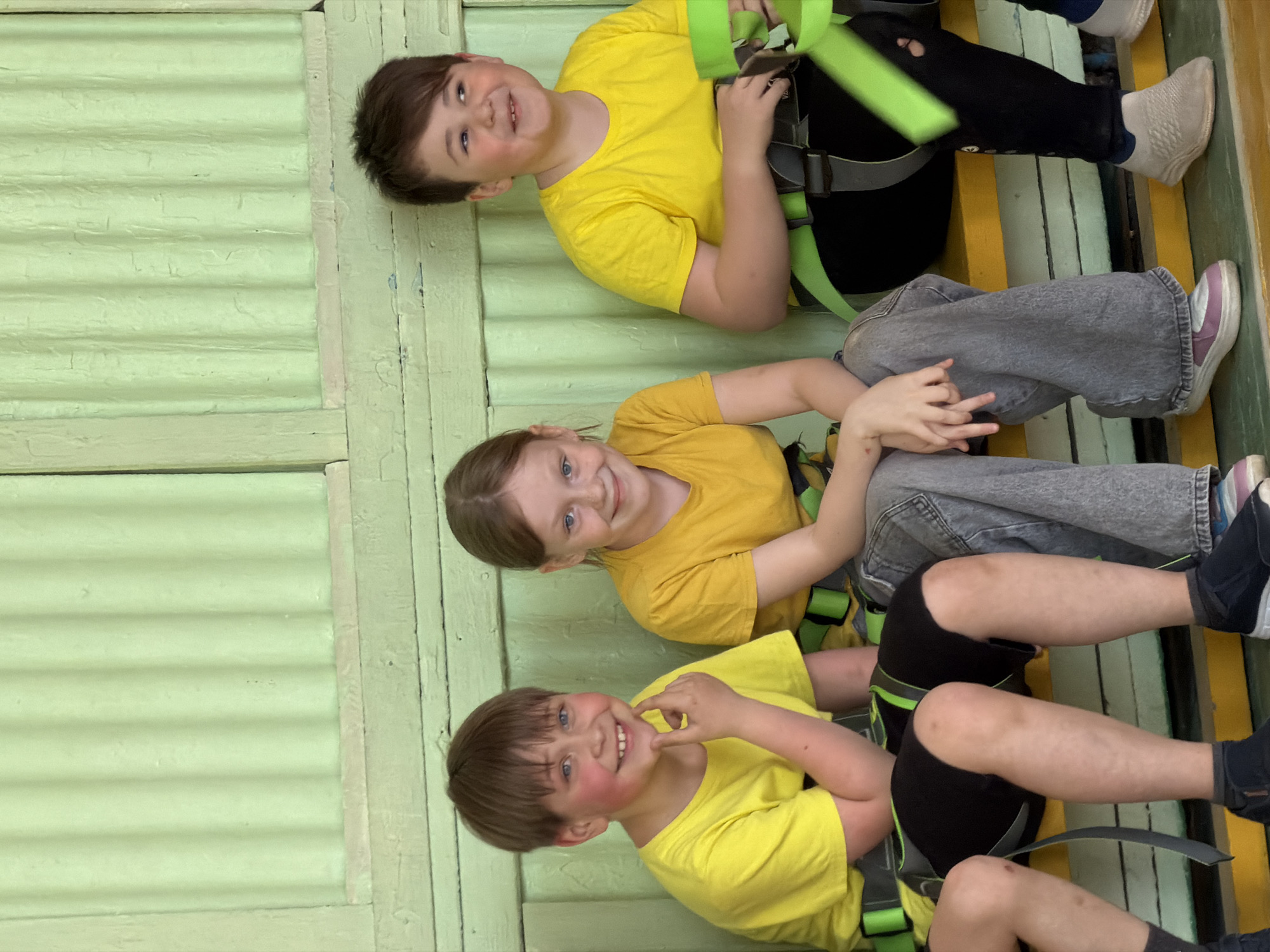 This image depicts four children participating in an activity at an indoor adventure park or climbing facility in Izhevsk, Russia. The image is oriented sideways, with the children appearing to be situated on a vertical structure, likely a wall or a climbing element.
All four children are wearing bright yellow t-shirts and green safety harnesses, typical for ropes courses or climbing activities. The children are seen from approximately the chest up, with their legs and the lower part of the structure extending out of frame at the bottom.
From top to bottom in the rotated image:
1. A boy with short, dark hair is visible from the head to the chest, smiling broadly at the camera. He is adjusting the green straps of his harness.
2. A girl with brown hair and blue eyes is smiling warmly at the camera. She is wearing a yellow t-shirt, gray jeans, and light-colored sneakers with purple details. Her hands are resting on her lap.
3. A boy with brown hair and blue eyes is also smiling directly at the camera, with one hand casually touching his chin. He wears a yellow t-shirt, black shorts, and dark shoes.
4. A fourth child is partially visible at the bottom of the frame, wearing a yellow t-shirt, black shorts, and a green harness.
The background features a light green, ribbed wall, suggesting painted wooden panels or siding. The climbing structure itself is made of wooden planks painted in sections of bright yellow and light green. The children's expressions are universally joyful, indicating a fun and engaging event. There is no visible text in the image. The setting appears to be indoors, making the time of day or external weather conditions indistinguishable.