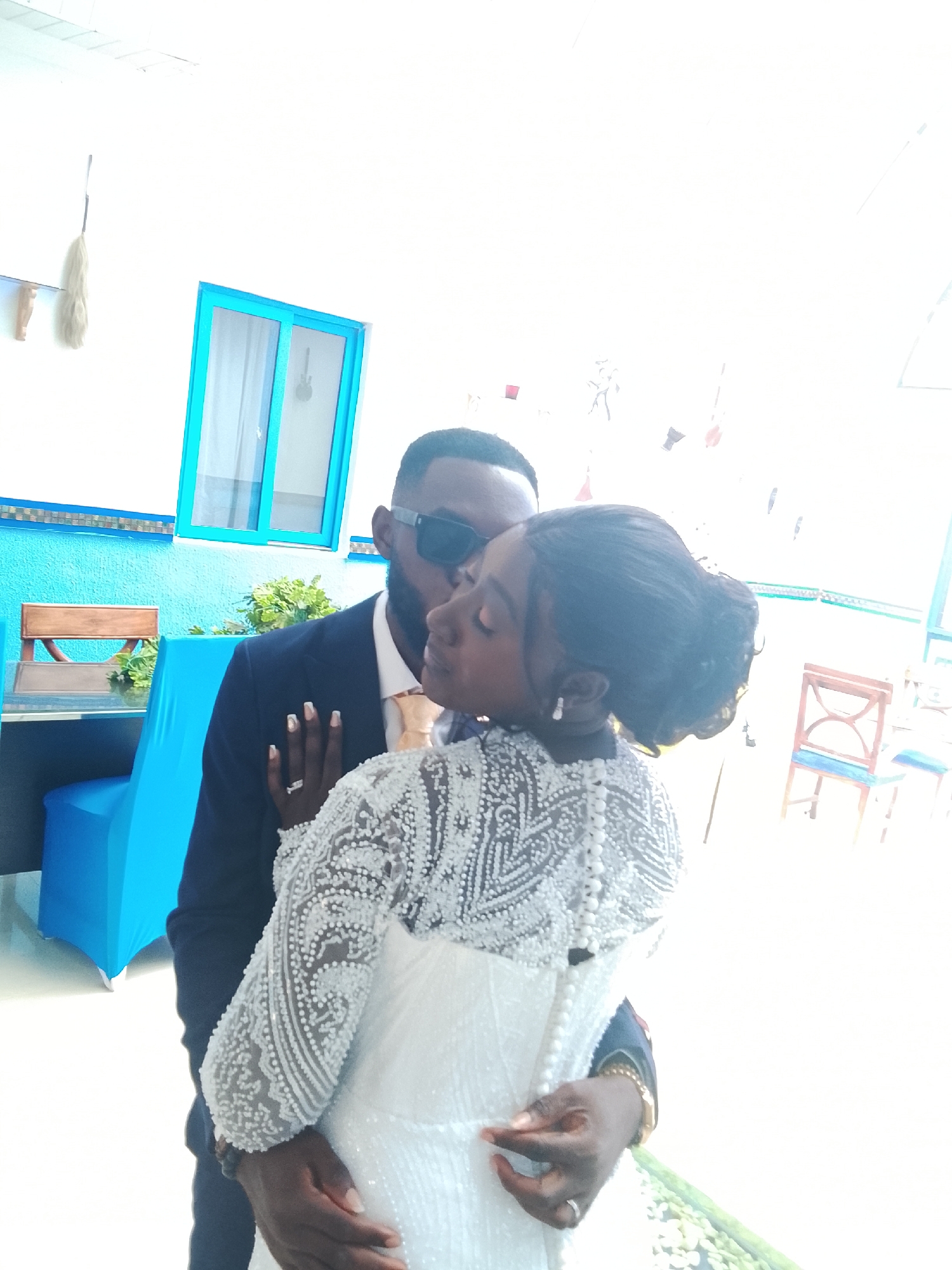 In an indoor setting in the city of Unknown, Unknown, a couple is captured in a close embrace. The man, wearing a dark suit, a light-colored tie, and sunglasses, has a beard and is kissing the woman on the side of her face. The woman is wearing a white, long-sleeved dress adorned with intricate, bead-like detailing. She has her hair styled up. Both are wearing rings on their fingers, indicating a possible wedding or engagement. The man's arm is wrapped around her waist. The background includes a blue-painted wall with a window trimmed in turquoise, and there's a table covered with a blue cloth. A couple of chairs are visible near the table. The overall lighting in the scene appears bright, with some areas appearing washed out.