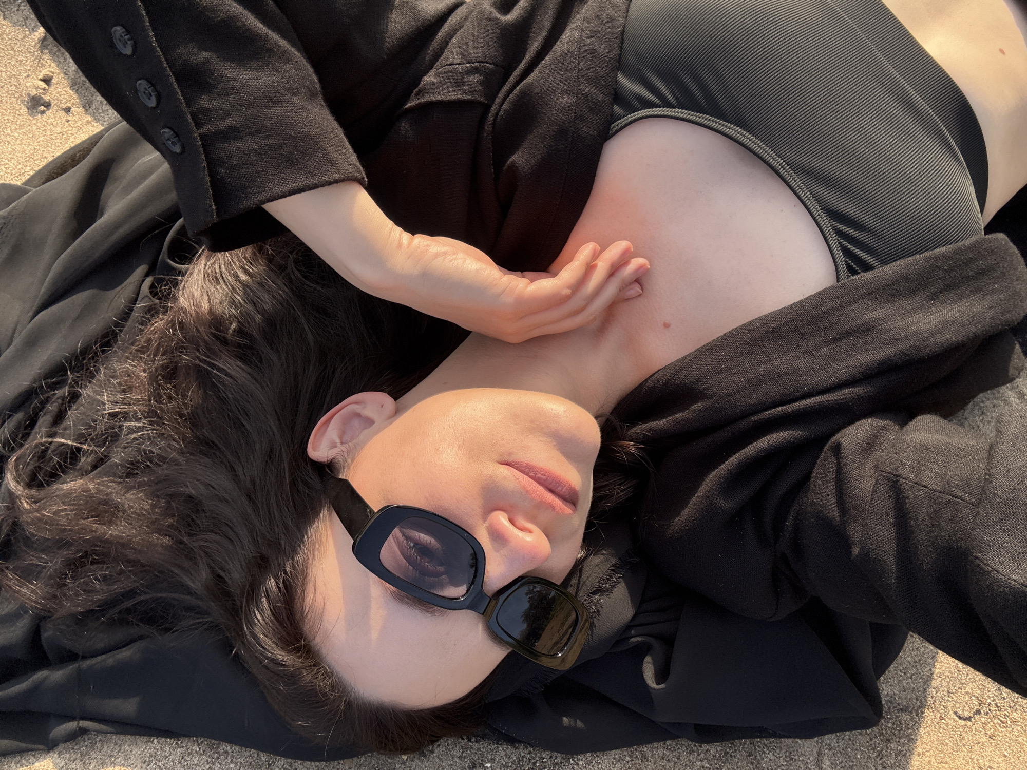 A young woman with dark hair and sunglasses is lying on her back on a sandy surface, possibly a beach. She is wearing a black jacket over a ribbed black top. Her left arm is resting on her chest, with her hand lightly touching her neck. The lighting suggests it is either sunrise or sunset, casting a warm glow on her skin. The background is out of focus, but the sandy texture is visible. There is no indication of the specific location in Chaykovsky, Russia.