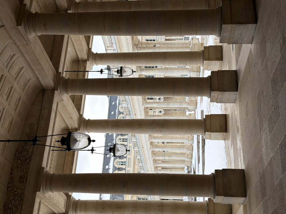 The image captures a detailed view of the snow-covered interior courtyard of the Palais Royal in Paris, France, observed from within a stone colonnaded portico. In the immediate foreground, light-colored fluted columns with square bases rise vertically, supporting a geometrically coffered stone ceiling adorned with intricate moldings. Three ornate black metal lanterns, featuring clear glass globes, hang from the ceiling at different depths, with the largest suspended centrally in the closest column bay. The floor of the portico is paved with large, irregularly shaped stone slabs, displaying variations in color and moisture.
Beyond the foreground columns, the central courtyard is uniformly blanketed in white snow, interrupted by some darker, possibly melted or disturbed patches. Several low-lying, reflective metallic spherical sculptures are partially visible within the snowy expanse. A parallel colonnade, composed of numerous identical stone columns, spans the width of the courtyard. In the background, multi-story classical buildings feature multiple windows, dark sloped roofs partially covered with snow, and distinctive dormer windows, all set against an overcast sky. The scene is devoid of any individuals or discernible human activity.