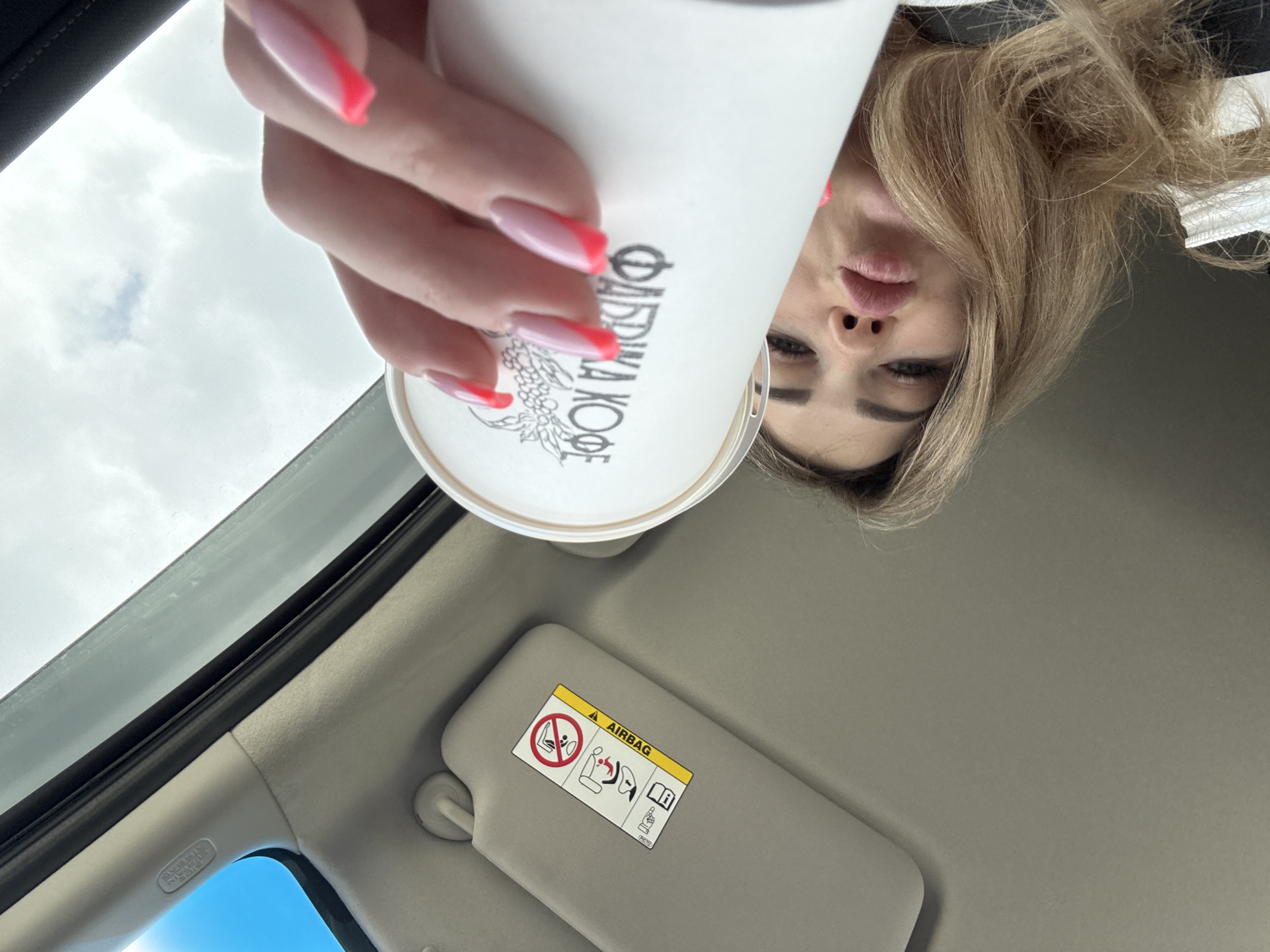 A woman with long blonde hair is holding a white paper coffee cup with pink-tipped nails. The cup has the Cyrillic text "ФАБРИКА КОФЕ" on it. She appears to be inside a car, based on the view from the window and the sun visor with an airbag warning. The sky is visible through the car window. The city is Unknown, Unknown.