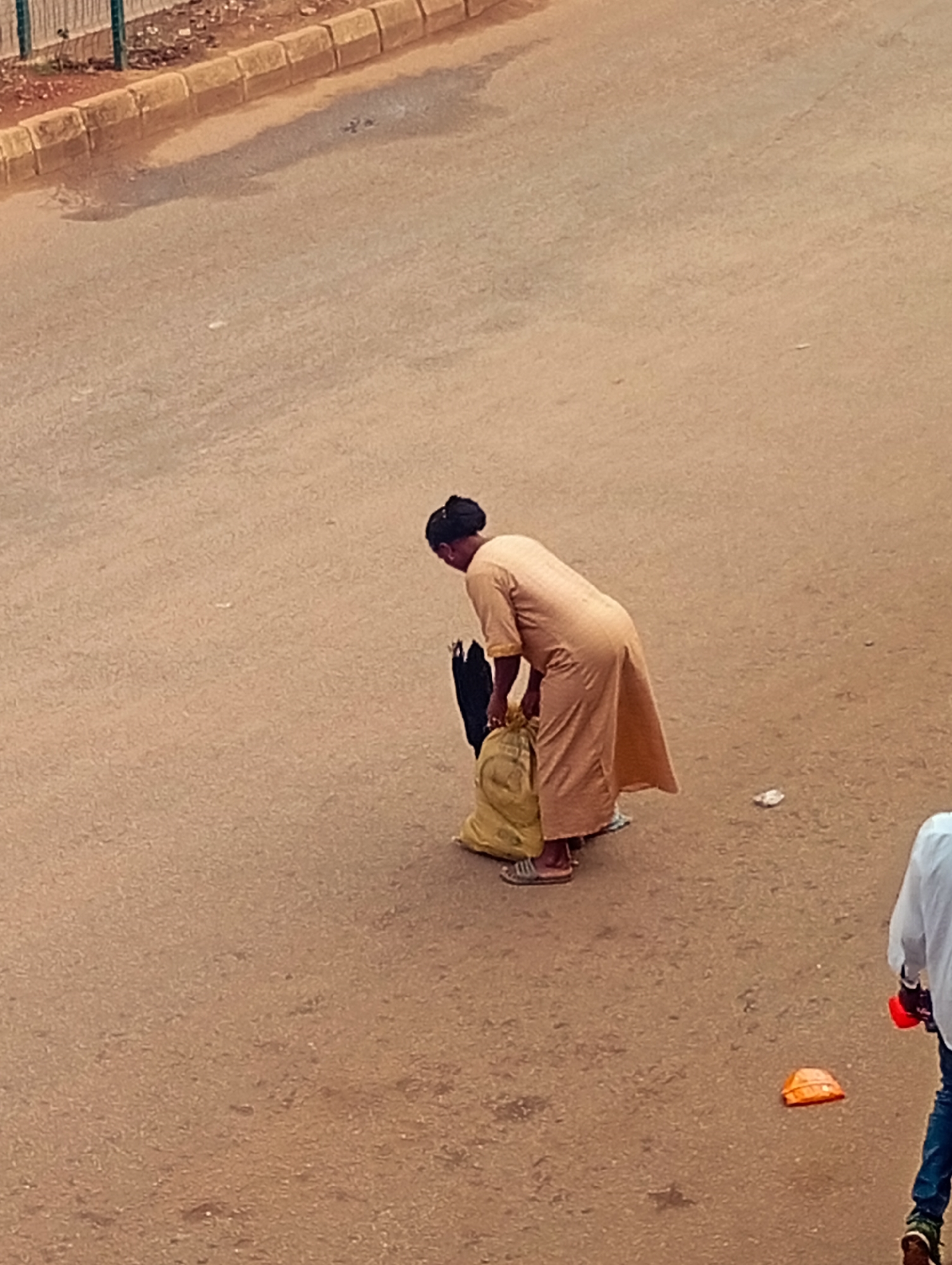 In Bukuru, Nigeria, a woman in a long, tan dress with her hair in a bun is bending down to adjust a large, yellow bag on the asphalt road. She is holding a black object, possibly an umbrella or another bag, in her left hand. To the right of the frame, a person wearing a white shirt and blue jeans is partially visible, walking away from the viewer. An orange object, resembling a plastic container or lid, lies on the road near the person in the white shirt. The scene takes place on a paved road with a curb and a metal fence in the background.