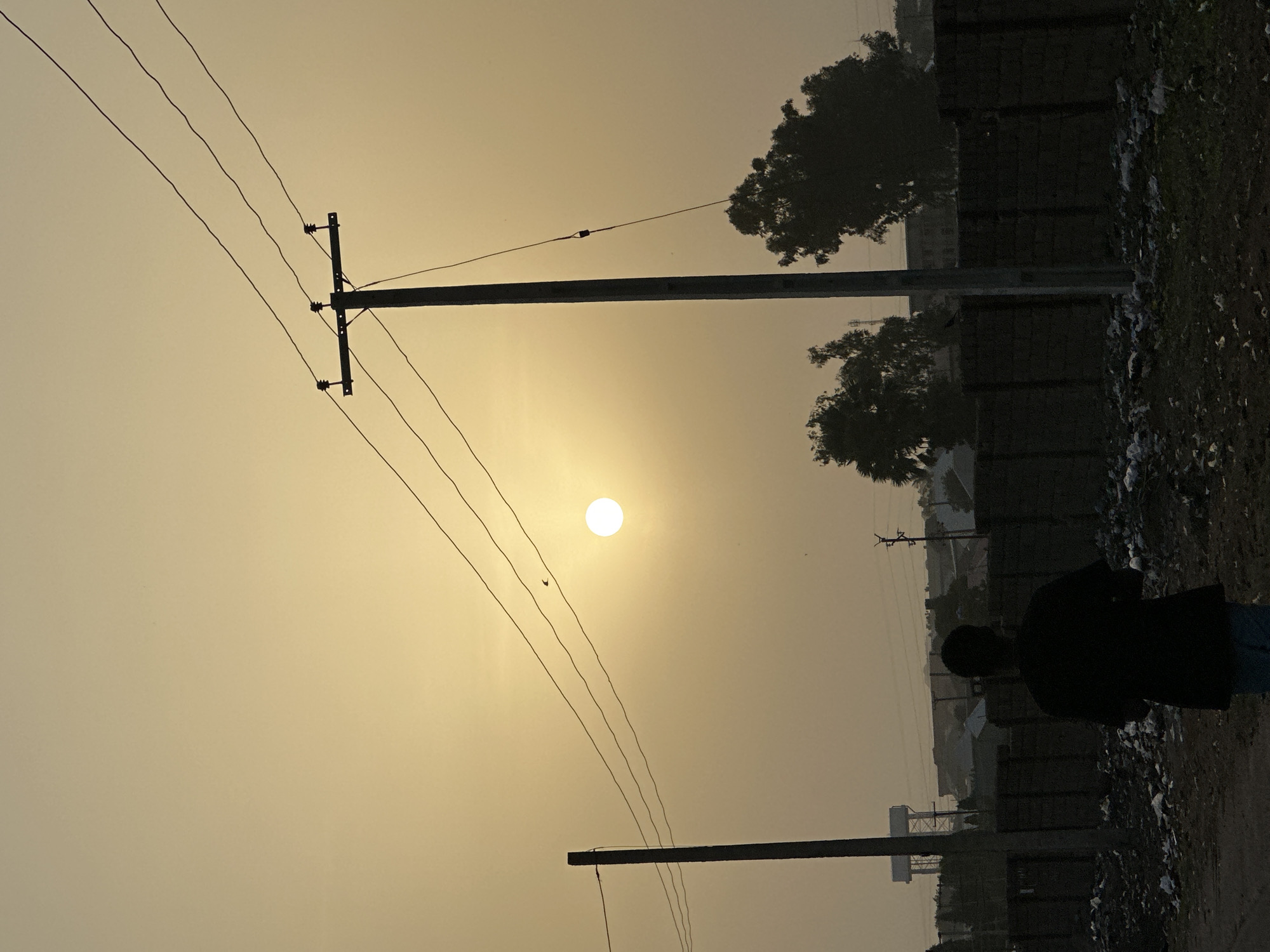 The image depicts an outdoor twilight scene, likely sunrise or sunset, in what appears to be a semi-urban or rural setting in Bukuru, Nigeria. The dominant feature is a bright, circular sun, appearing almost white, positioned centrally in a hazy, warm golden-yellow sky. This strong backlighting creates prominent silhouettes of all foreground and mid-ground elements.
On the right side of the frame, a lone person stands facing away from the camera, looking towards the left horizon. They are entirely silhouetted, wearing a dark top, with a subtle hint of blue on what might be their trousers. The ground beneath them is unpaved, appearing as dirt or gravel, with scattered debris or litter, predominantly white fragments, visible.
Two utility poles are prominent. A taller pole with multiple cross-arms and several thin power lines extends from the left side of the frame, stretching horizontally across the sky towards the right. Another shorter, thicker pole is partially visible in the lower right foreground. Numerous power lines crisscross the hazy sky, adding to the visual texture.
In the mid-ground, behind the person, is a dark, undefined structure that resembles a long wall or a fence, possibly part of a building. Silhouetted trees with dense foliage are visible behind this structure and also in the upper right quadrant of the image. Further in the distance, faint outlines of more buildings and what appears to be a water tower or similar structure are discernible, softened by the atmospheric haze.
The overall mood is quiet and contemplative, emphasized by the solitary figure and the soft, diffused light of twilight. There is no visible text in the image.