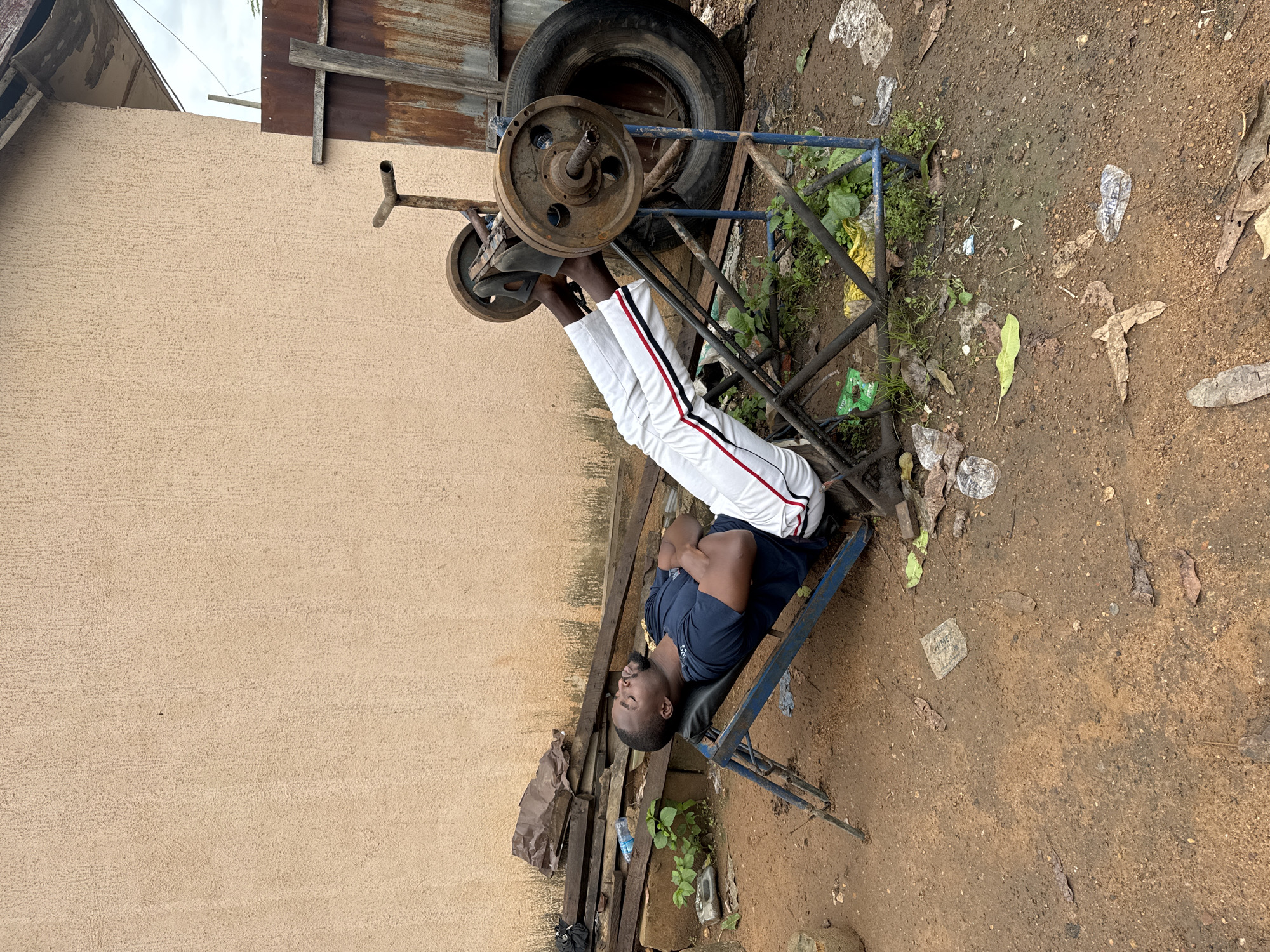 A man is lying on his back on a weight bench, his feet hooked under a barbell. He is wearing white pants with red and black stripes down the sides, and a dark blue t-shirt. The weight bench is made of blue metal and appears to be old and rusty. It is situated outdoors on a dirt ground, with debris scattered around. In the background, there is a textured light brown wall, and a rusty corrugated metal structure. Above the man's head, a large tire is visible. The man's eyes are closed, and his arms are crossed over his chest, suggesting he is resting or taking a break from his workout. The overall scene depicts an improvised outdoor gym, possibly in a rural or less developed area, given the condition of the equipment and the surrounding environment. The lighting suggests it is daytime, possibly overcast.