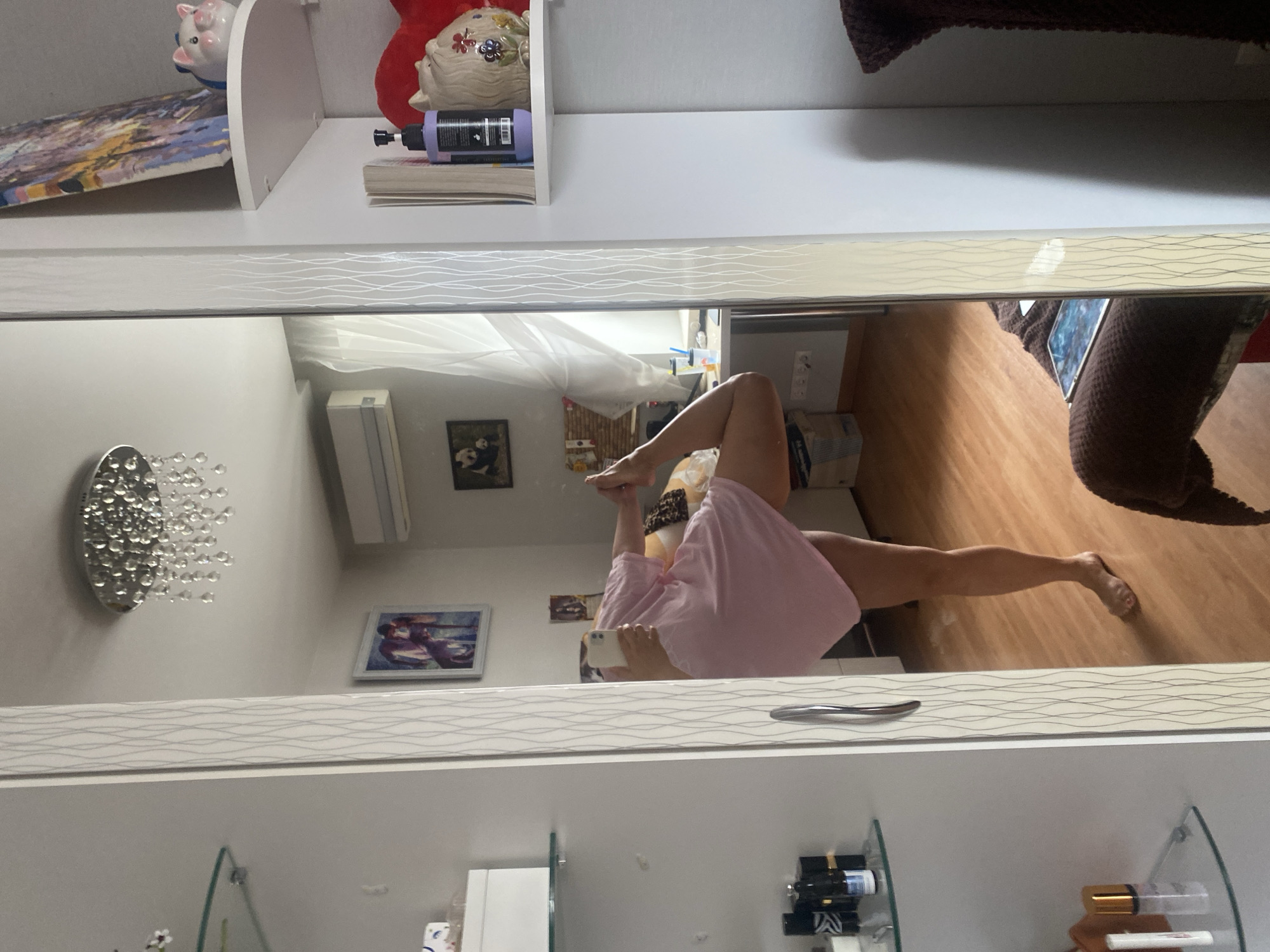 The image shows a person in a pink t-shirt and leopard-print shorts, captured in a mirror reflection. The person is performing a yoga pose, with one leg extended upwards and the other bent. They are holding a smartphone in their hand, seemingly taking a selfie. The setting appears to be indoors, likely a home, with wooden flooring visible in the reflection. In the background, there are shelves with various items, including decorative pigs, a painting, and bottles of products. There is also a framed picture on the wall and an air conditioning unit. The overall mood is casual and relaxed. The reflection also shows a portion of what appears to be a closet or cabinet with decorative wavy lines. The image is oriented such that the subjects are upside down, indicating it was taken with the camera rotated.