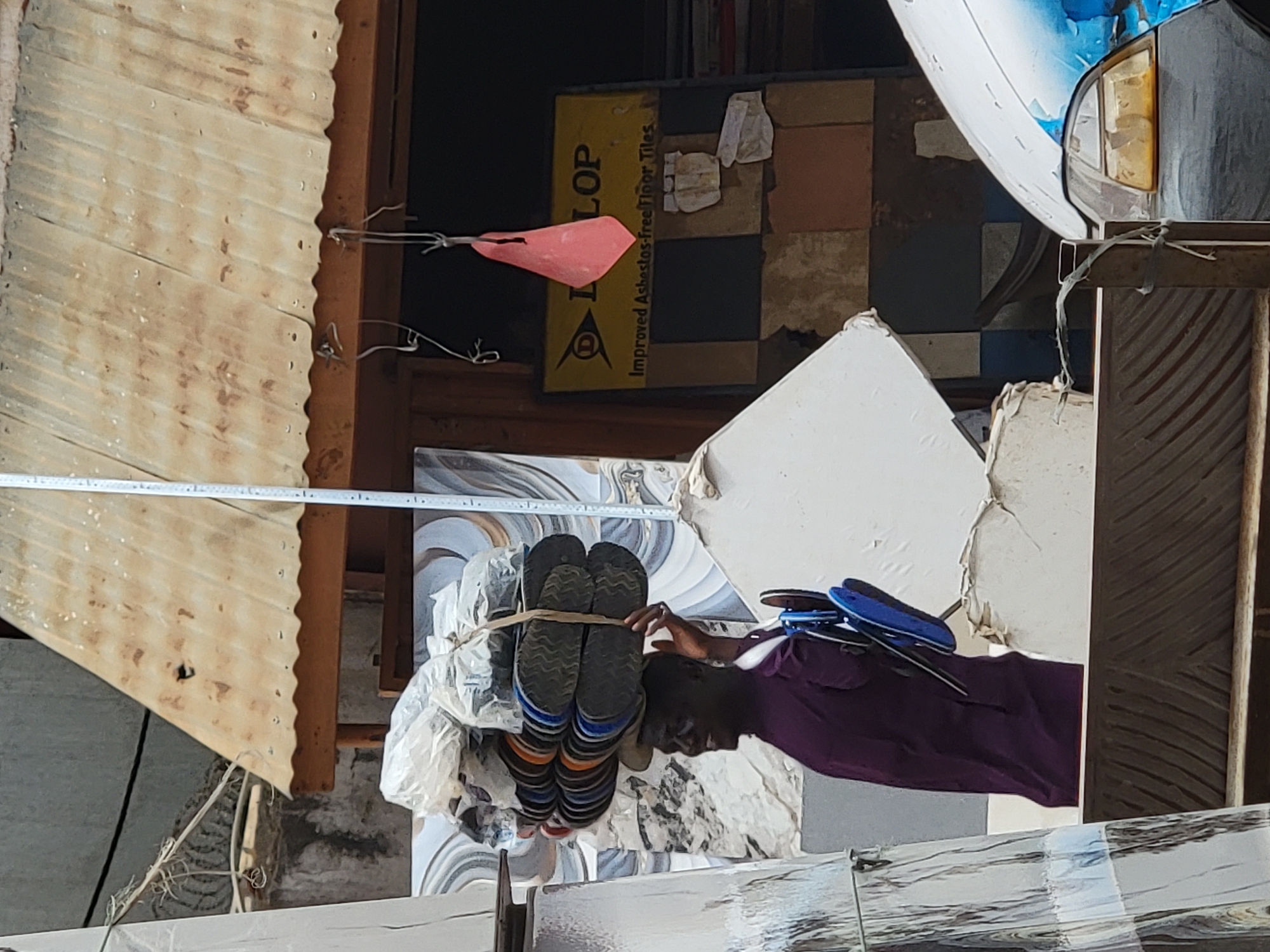 This image, originally rotated ninety degrees clockwise, captures a vibrant market or vendor stall scene in Maiburiji, Nigeria, during daylight hours. The central figure is a man with dark skin, dressed in a purple shirt, who appears to be organizing or handling numerous pairs of sandals or flip-flops. These sandals, featuring various color combinations including black, blue, and orange, are bound together with string and prominently displayed.
The setting is characterized by a functional and somewhat cluttered environment, typical of a busy marketplace. To the left, a corrugated metal sheet suggests a makeshift structure or roof. A long measuring tape extends across the upper mid-ground. In the background, a large yellow sign is partially visible, bearing the text "LOP" (likely part of 'DUNLOP') and the phrase "Improved Asbestos-free Floor Tiles," along with the distinctive Dunlop logo. Below this sign, different patterned tiles are arranged, indicating the sale of construction or home improvement materials.
Other notable objects include a red, conical object hanging near the sign and the front section of a vehicle, possibly a car or commercial tricycle, with a headlight and peeling blue paint, positioned on the right. The scene encapsulates the everyday commercial activity of a local Nigerian market, with the man focused on his work amidst a variety of goods and materials.