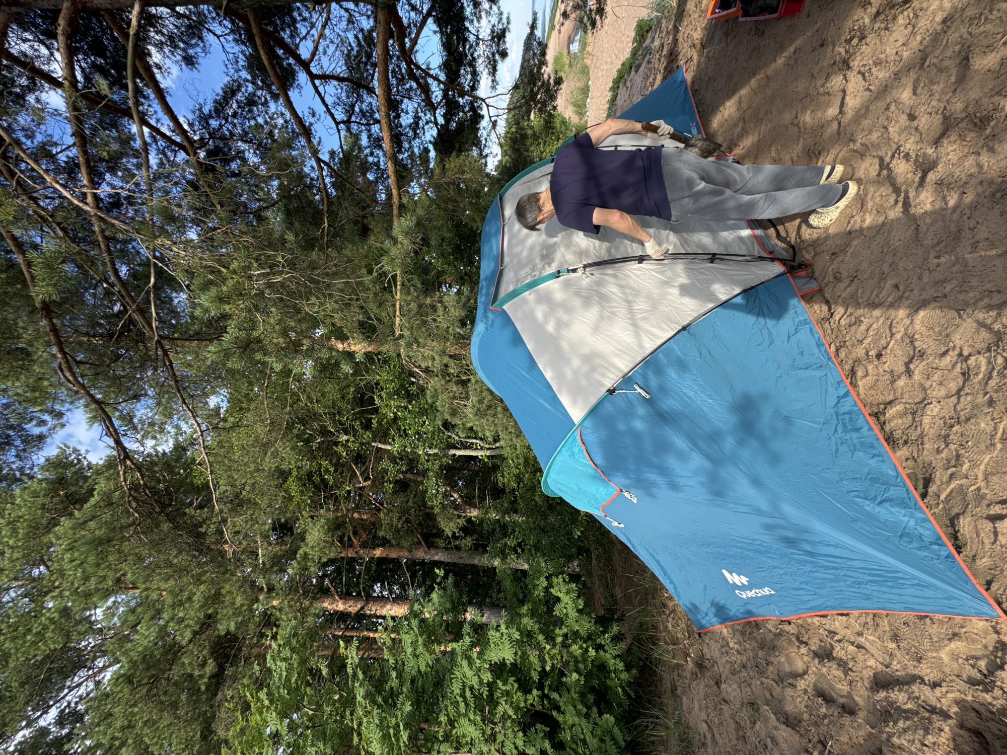 The image depicts a man in a dark blue t-shirt and grey pants wearing white gloves and light-colored clogs. He is standing near a partially set-up blue and white tent on a sandy area, holding a small shovel. The tent is situated in a natural environment with trees and foliage in the background. The sandy area appears to be near a body of water, possibly a beach. The sky is visible and appears to be blue with some clouds. This scene is located in the city of Zelenaya Roshcha, Russia.