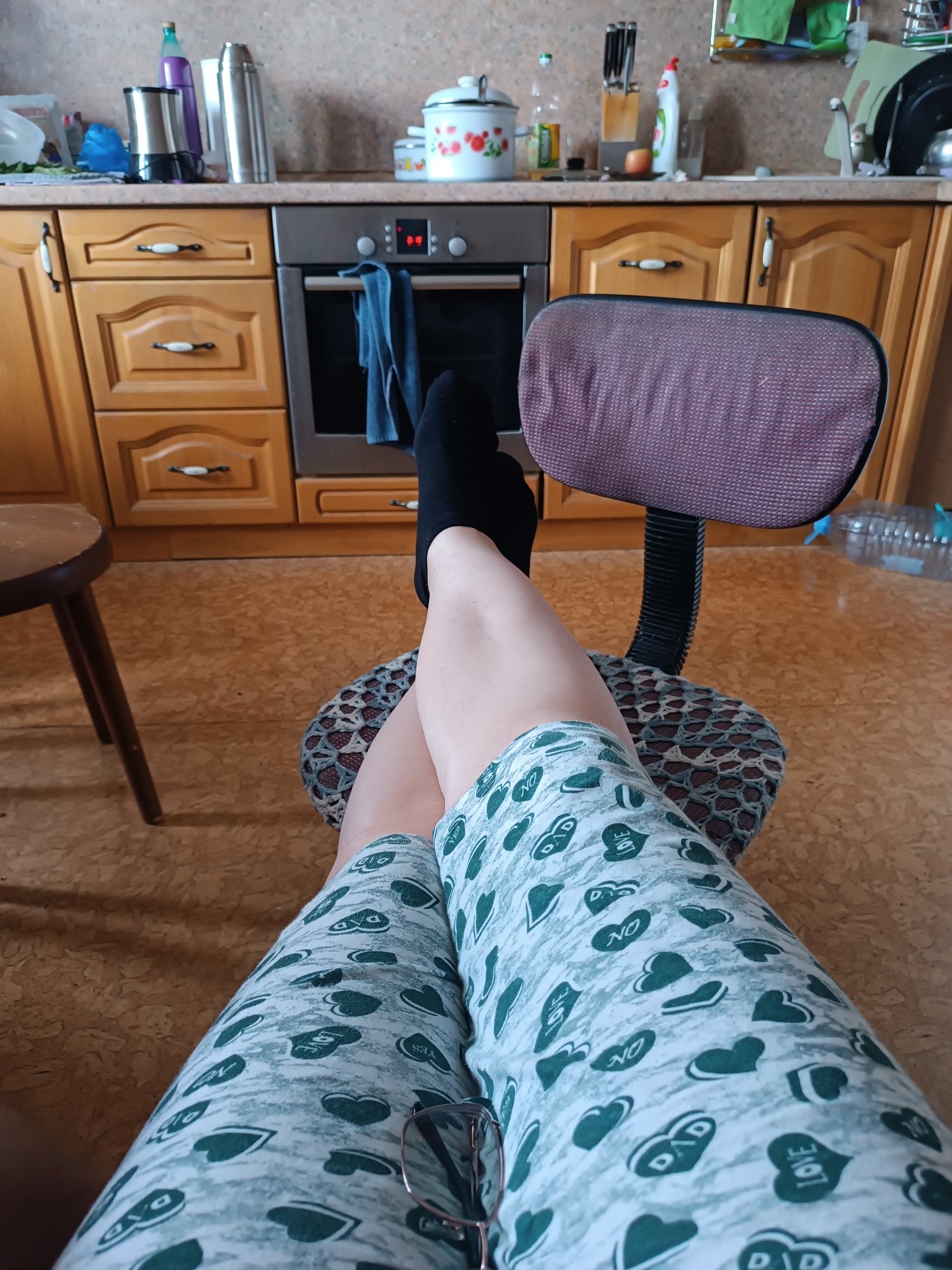 The image shows a person's legs and feet in a kitchen environment. The person is sitting in a chair with a patterned seat and back, wearing black socks and patterned shorts with green hearts and words. The kitchen features wooden cabinets, a stove, and a countertop with various items, including appliances, bottles, and containers. A wooden stool is visible to the left of the person. The floor is a light brown, cork-like material. Glasses are resting on the person's leg.