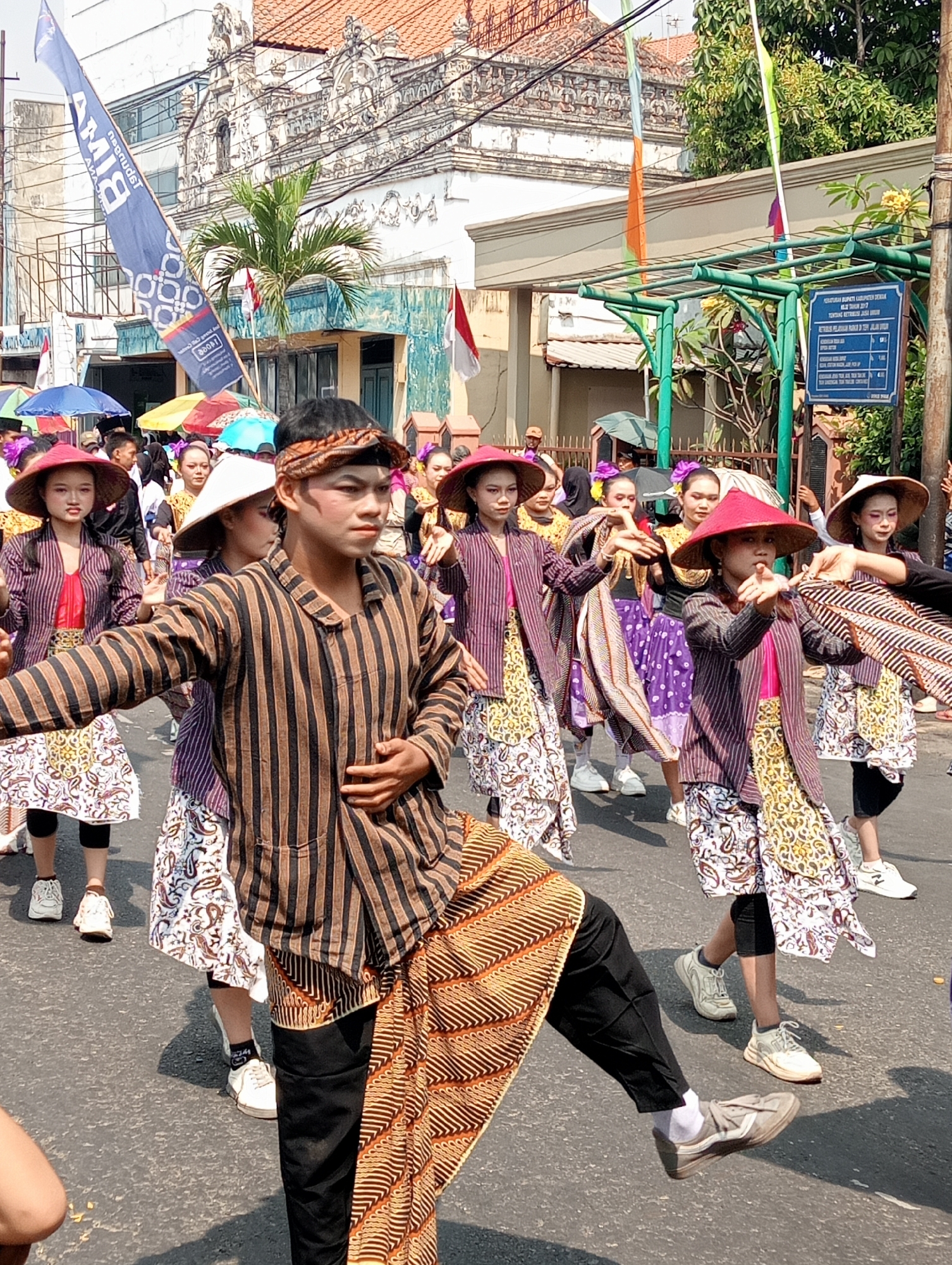A group of people, primarily young women, are participating in a parade on a paved street in Bintoro, Indonesia. The parade appears to be a cultural event, with participants dressed in traditional clothing.
The central figure is a young man with theatrical makeup, wearing a brown and black striped tunic over a sarong with an orange and brown pattern. He has a patterned cloth headband and is performing a dance move with his right leg extended.
To his left, several young women are dressed in similar striped tunics, layered over brightly colored tops and patterned skirts. Many are wearing wide-brimmed hats, some straw and some with red fabric. They are holding decorative fabrics or cloths in their hands, suggesting they are part of a choreographed performance.
In the background, more participants are visible, along with colorful umbrellas and flags. The buildings in the background suggest an urban setting, with ornate architecture. A blue banner with white text and circular patterns is visible on the left, and a sign with Indonesian text is mounted on a green structure on the right. The lighting indicates it is daytime, likely sunny, casting shadows on the street.