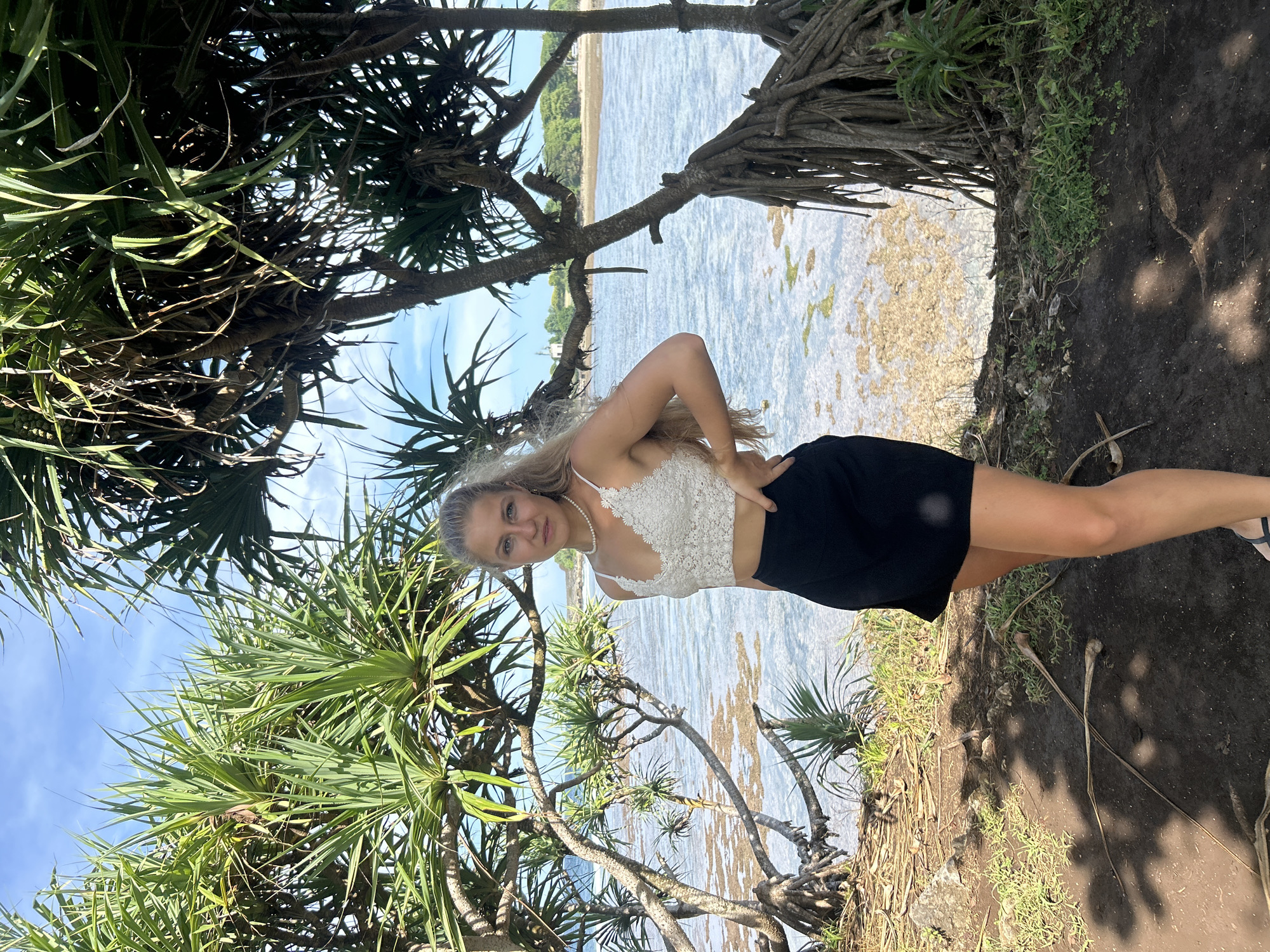 The photograph depicts a young woman posing against a backdrop of tropical flora and an ocean view, likely taken in the vicinity of Kuta Selatan, Indonesia. The woman has blonde hair and is wearing a white lace top and a black skirt. She is standing on a dirt path, and behind her is a body of water with visible coral or rock formations beneath the surface. The scene is framed by trees with long, spiky leaves, which are typical of tropical coastal vegetation. The sky is visible in the upper portion of the image, suggesting it is daytime.