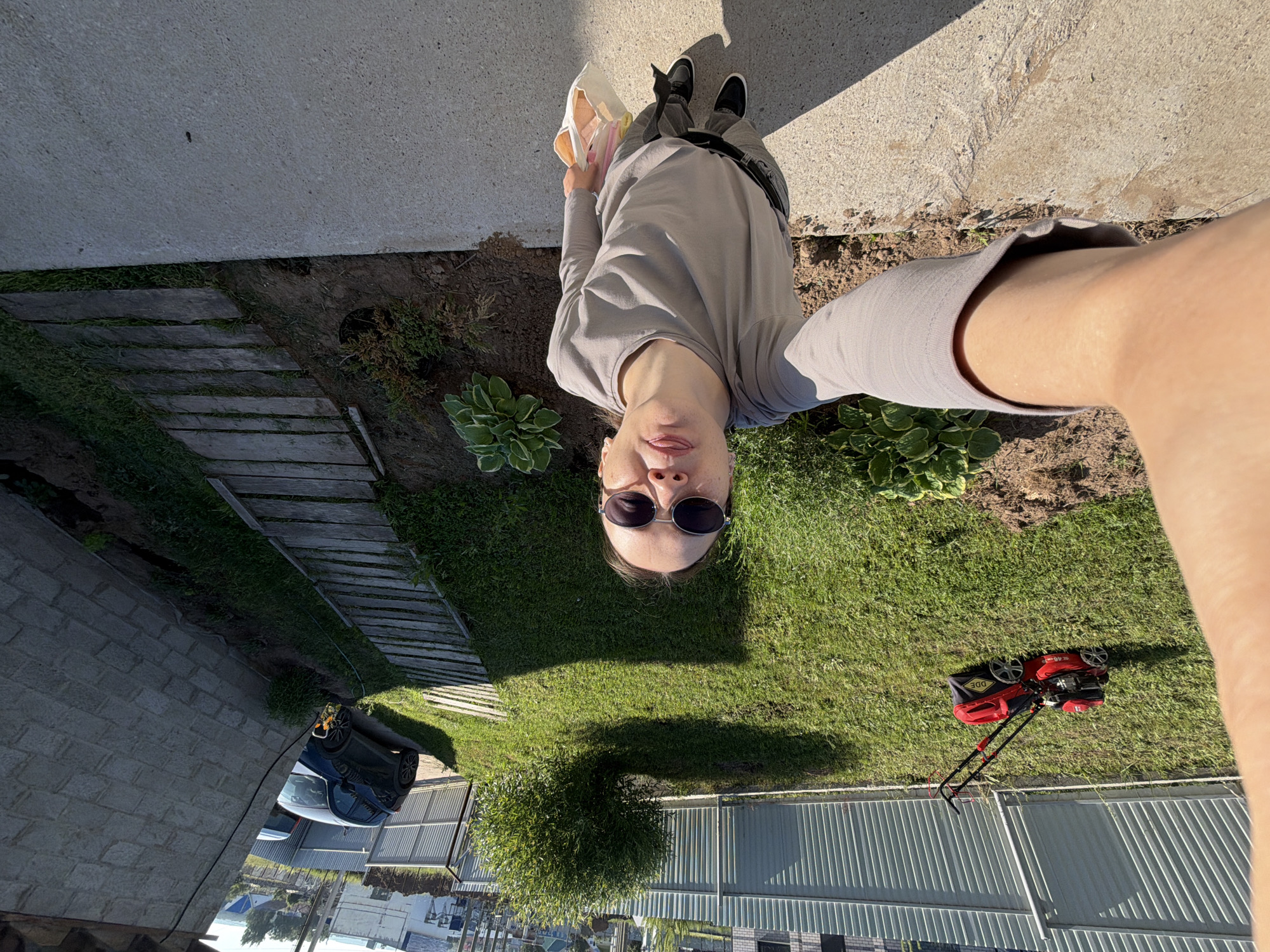 The image depicts a full-body, wide-angle selfie taken outdoors in what appears to be a residential backyard in Chaikovsky, Russia. The subject, a person wearing a grey long-sleeved shirt, grey pants, black sneakers, and sunglasses, is standing on a concrete path, looking up towards the camera with their tongue sticking out. They are holding a light-colored tote bag.
The background includes a green lawn, a red lawnmower, a dense green bush, a wooden plank walkway, a light-colored brick building, a metal fence, and two cars parked in the distance. The scene suggests a sunny day with shadows cast on the ground. The overall impression is a candid shot taken in a suburban or rural setting.
