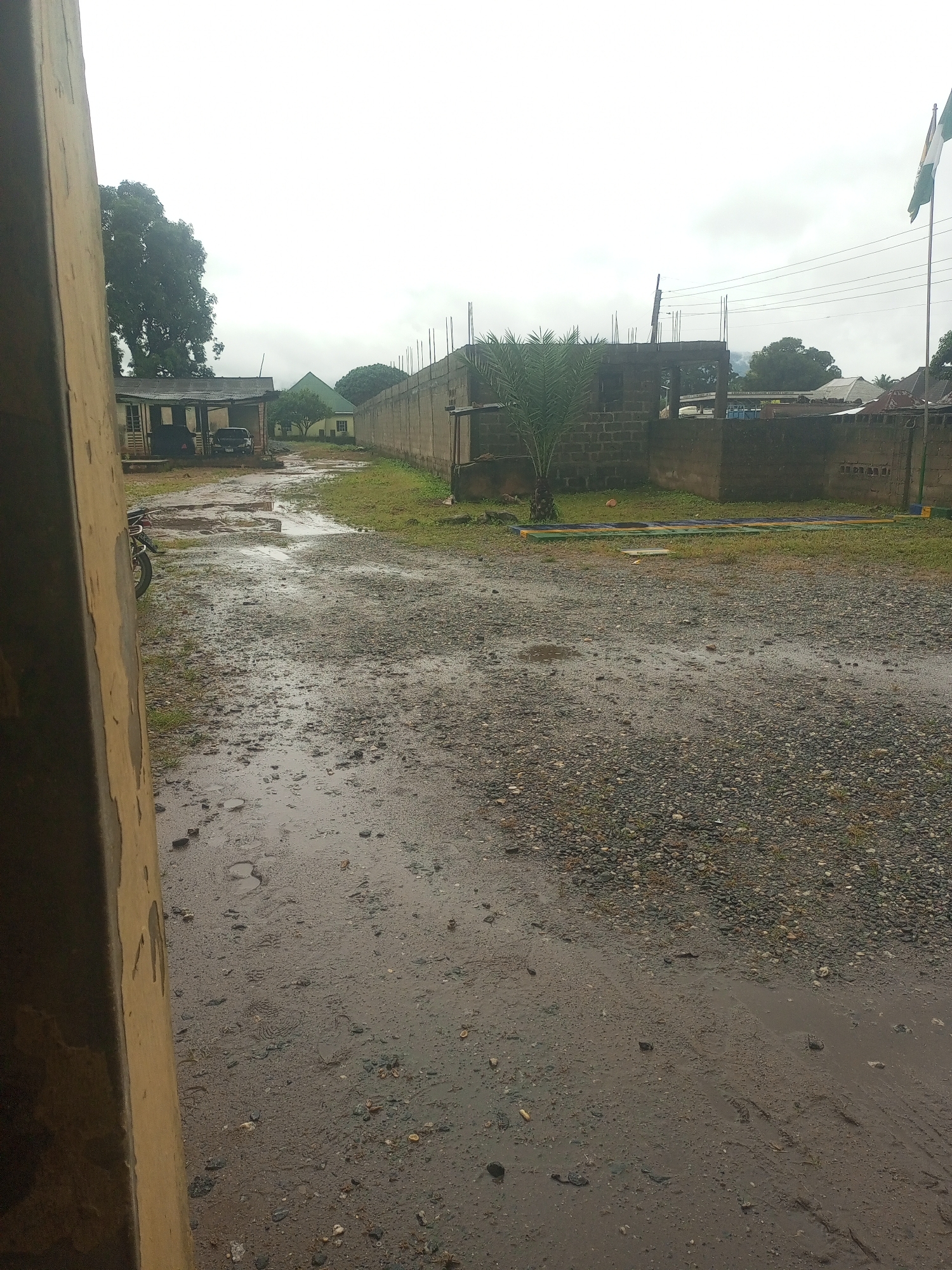 The image depicts an outdoor scene, likely in Serti, Nigeria, with a gravelly, wet pathway in the foreground. The ground is uneven, showing evidence of puddles and tire tracks, suggesting recent rainfall. To the left, a portion of a weathered, light-colored structure, possibly a wall or a building facade, occupies the edge of the frame. Further back, a single motorcycle is parked, its front wheel visible. Across the pathway, a low-lying building with a corrugated iron roof stands partially obscured by foliage. In the background, a construction site is evident, with concrete blocks forming the walls of an unfinished structure and rebar protruding from the top. Beyond the construction, a long concrete wall extends across the scene, marking a boundary. A flag, predominantly green and white, flies from a flagpole on the right side of the image, against a cloudy sky. The environment appears somewhat unkempt, with scattered debris and overgrown grass in some areas. The overall impression is one of a rural or semi-urban setting experiencing inclement weather.