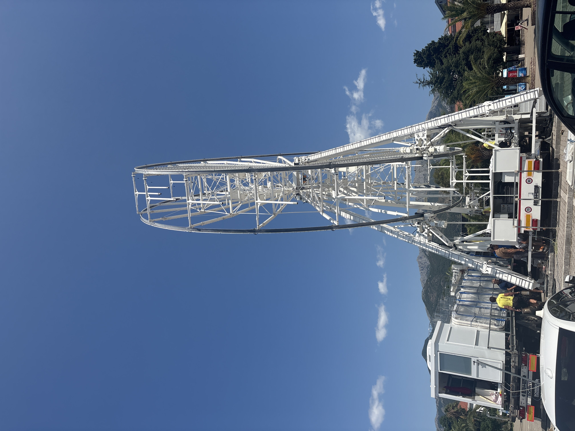 The image shows a partially constructed Ferris wheel against a clear blue sky. The Ferris wheel structure is white and made of metal beams. Workers are visible on and around a white truck positioned next to the Ferris wheel, seemingly engaged in construction or assembly. One worker in a yellow shirt is working near a white cabin-like structure on the truck. Another worker in a brown shirt is higher up on a ladder attached to the Ferris wheel structure. The scene appears to be outdoors, with palm trees and other greenery visible in the background, along with mountains in the distance. The bright sunlight suggests it is daytime. The overall setting suggests a public space or a fairground in a warm climate, possibly near a coastal area given the presence of palm trees. There is no clear indication of emotion, but the activity suggests a process of construction or setup. Visible text on the truck includes "SCHMITZ CARGO" and "CARAVANS" on the rear, along with some circular red and white signs with numbers that are difficult to read clearly. The image is oriented vertically.
