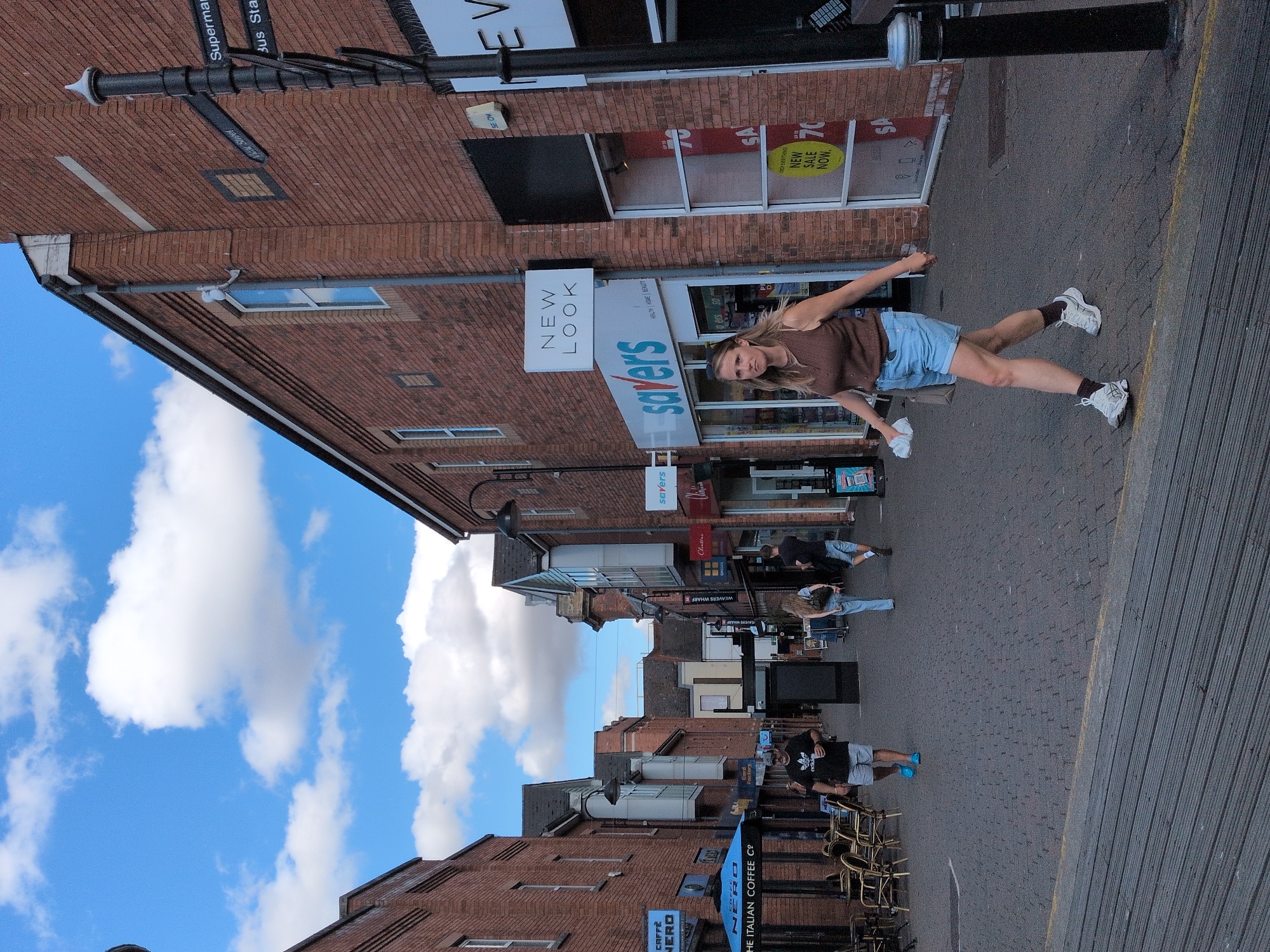 The image captures a street scene in Wyre Forest, United Kingdom, under a partly cloudy sky. The street is paved with dark cobblestones and bordered by traditional brick buildings lining both sides. Several people are visible walking along the pedestrianized street.
On the left side of the street, there is a Caffè Nero establishment with outdoor seating and a blue umbrella. Further down, a man wearing an Adidas t-shirt and grey shorts, along with bright blue flip-flops, is walking towards the viewer.
On the right side of the street, a woman is walking towards the viewer, wearing a brown sleeveless top, light blue denim shorts, white sneakers, and dark socks. She is holding a white tissue or cloth in her right hand and a light-colored bag in her left hand. Next to her, the storefronts of "New Look" and "Savers" are visible. The "Savers" store has a prominent sign indicating a "NEW SALE NOW."
Further down the street, two other individuals are walking away from the viewer. One is a woman with long blonde hair wearing a light-colored top and blue jeans, carrying a backpack. The other is a man in a black t-shirt and light blue shorts.
A black lamppost with street signs indicating "Supermarket," "View Street," and "Bus Station" stands on the right side of the street. The overall atmosphere suggests a typical day in a UK town center with shops and shoppers.