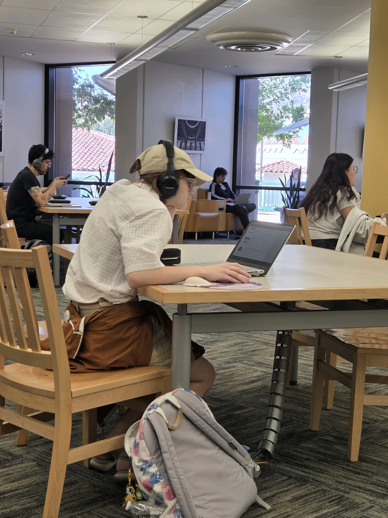 The image shows a group of people studying in a library in Glendale, USA. In the foreground, a person wearing headphones and a cap is seated at a table, using a laptop. To their left, another person with headphones is looking at their phone. In the background, two more individuals are seated at tables, one using a laptop and the other with their back to the camera. The library has large windows that offer a view of trees and buildings. The overall environment appears to be a quiet and studious setting.