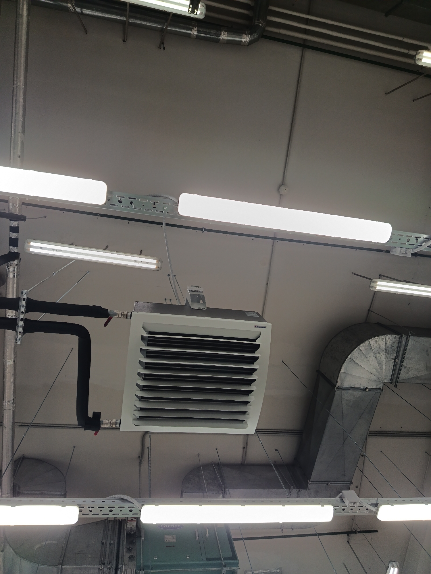 The image depicts a section of a building's interior, specifically the ceiling area, possibly in Moscow, Russia. Prominent elements include a white ceiling-mounted heater with horizontal vents, connected to black insulated pipes with red valves. Overhead fluorescent lights are interspersed with metal ductwork and pipework, attached to the ceiling with metal supports and wires. The overall impression is of a utility space with exposed mechanical and electrical systems.