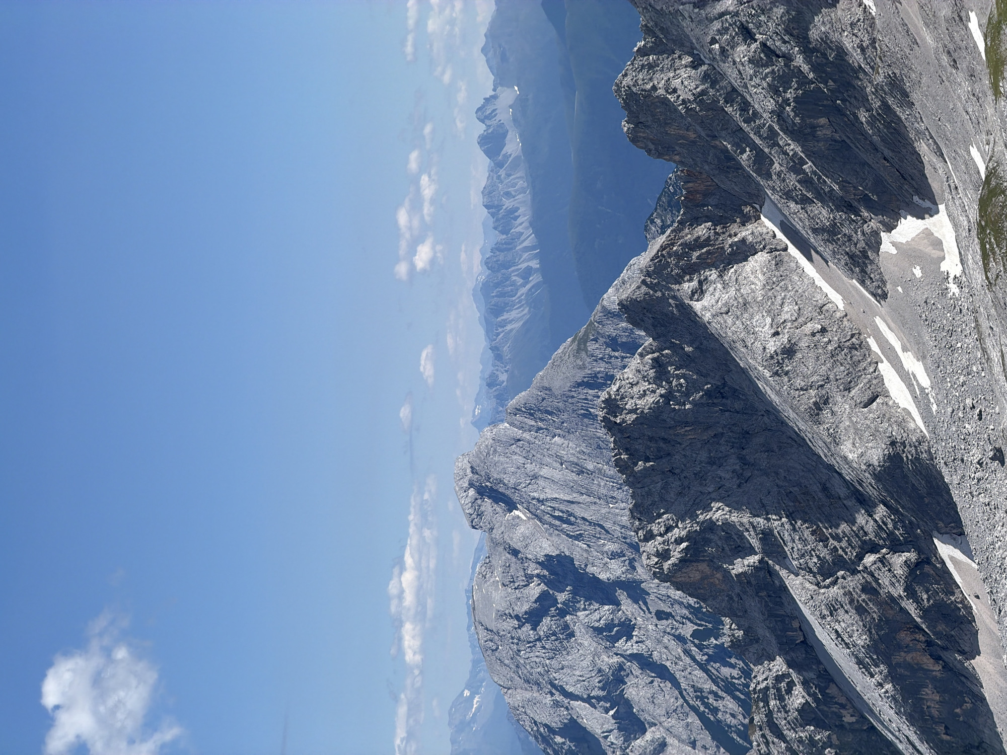 The image depicts a panoramic view of rugged, gray mountain peaks under a clear blue sky with scattered white clouds. The foreground is dominated by steep, rocky slopes with patches of snow. In the mid-ground, larger, more massive mountain formations rise, their surfaces textured by layers of rock and scree. In the distance, a series of progressively bluer mountain ranges recede into the horizon, indicating depth and a vast mountainous landscape. The scene is devoid of human presence or any man-made structures, focusing solely on the natural grandeur of the alpine environment. This vista is likely from a high vantage point overlooking the Wetterstein Mountains near Garmisch-Partenkirchen, Germany, a region renowned for its dramatic alpine scenery.