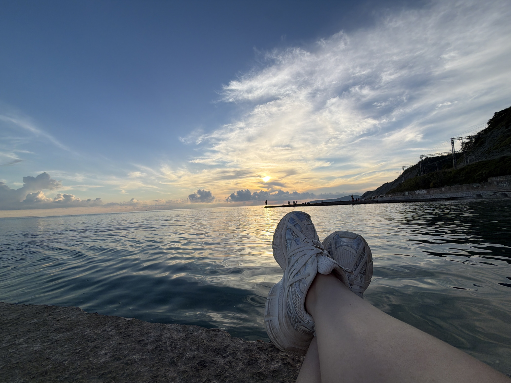 A low-angle, wide shot captures a serene sunset over the Black Sea in Dederkoy, Russia. The foreground features a person's legs and feet, clad in light-colored athletic shoes, resting on a rough, gray concrete surface. The person's skin is visible on their left thigh and shin. The sea, a deep blue, reflects the warm hues of the setting sun, with gentle ripples distorting the light. In the middle ground, a long pier or breakwater extends into the water, with a few silhouetted figures standing on it. The horizon is marked by a distant, dark shoreline and a steep, tree-covered hill on the right, upon which electrical pylons and wires are visible. The sky above is a gradient of blue, transitioning to lighter shades near the horizon, with wispy white clouds scattered across it, illuminated by the setting sun. The overall atmosphere is tranquil and picturesque.