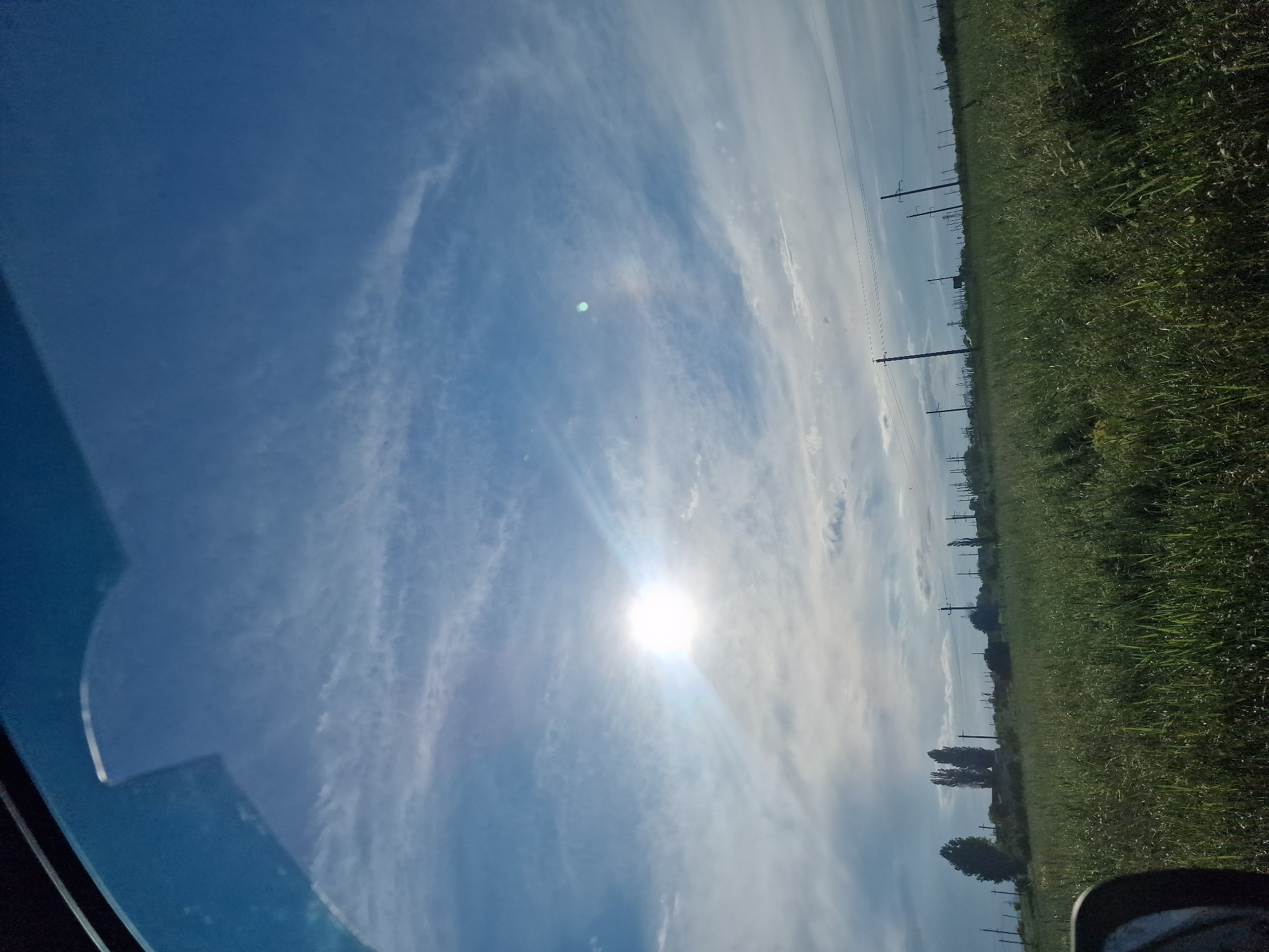 The image shows a rural landscape with a large, bright sun in a partly cloudy blue sky. In the foreground, there is a field of green grass and vegetation. In the mid-ground, there are several power lines supported by poles, and scattered trees. The sky is dominated by wispy white clouds. The scene appears to be taken from inside a vehicle, as a portion of a car's side mirror or window frame is visible in the lower left corner. The setting is consistent with the outskirts of a rural area like Kursavka, Russia, where agricultural land and infrastructure like power lines are common. There are no people or animals visible in the image.