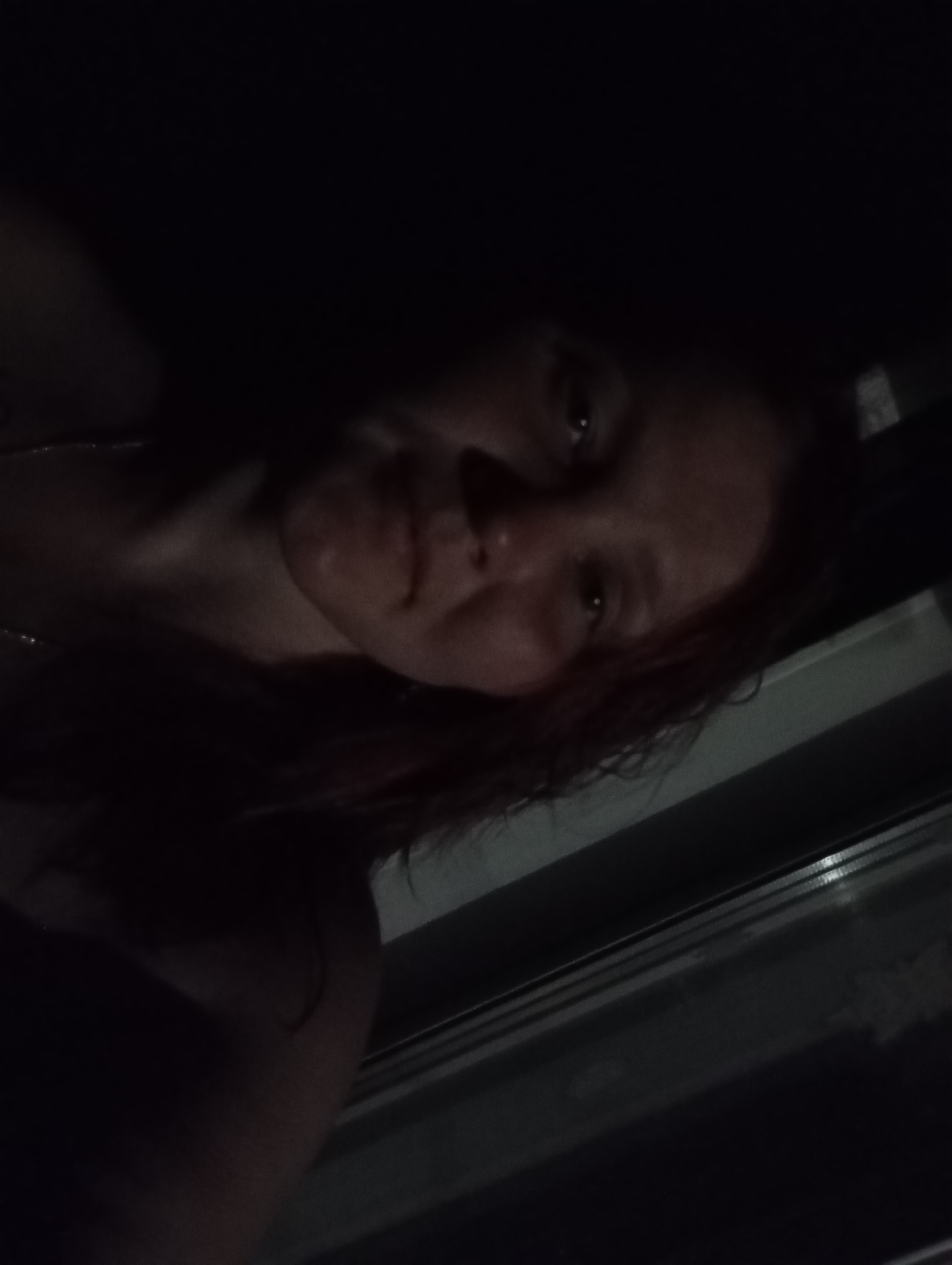 A woman is centered in the image, her face visible although the lighting is low. She has shoulder-length hair. The background is dark, suggesting a nighttime or dimly lit indoor environment. A window frame is partially visible in the lower right corner. The location is Unknown, Unknown.