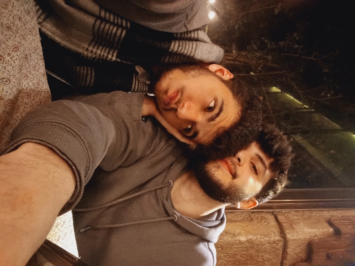Two young men pose closely in a selfie. The man on the right wears a gray hoodie and has a dark beard and curly hair. The man on the left has similar facial hair and dark eyes; he rests his head on the other man's shoulder. A gray, patterned blanket is behind the first man and a window reflecting outdoor greenery is visible behind the second man.