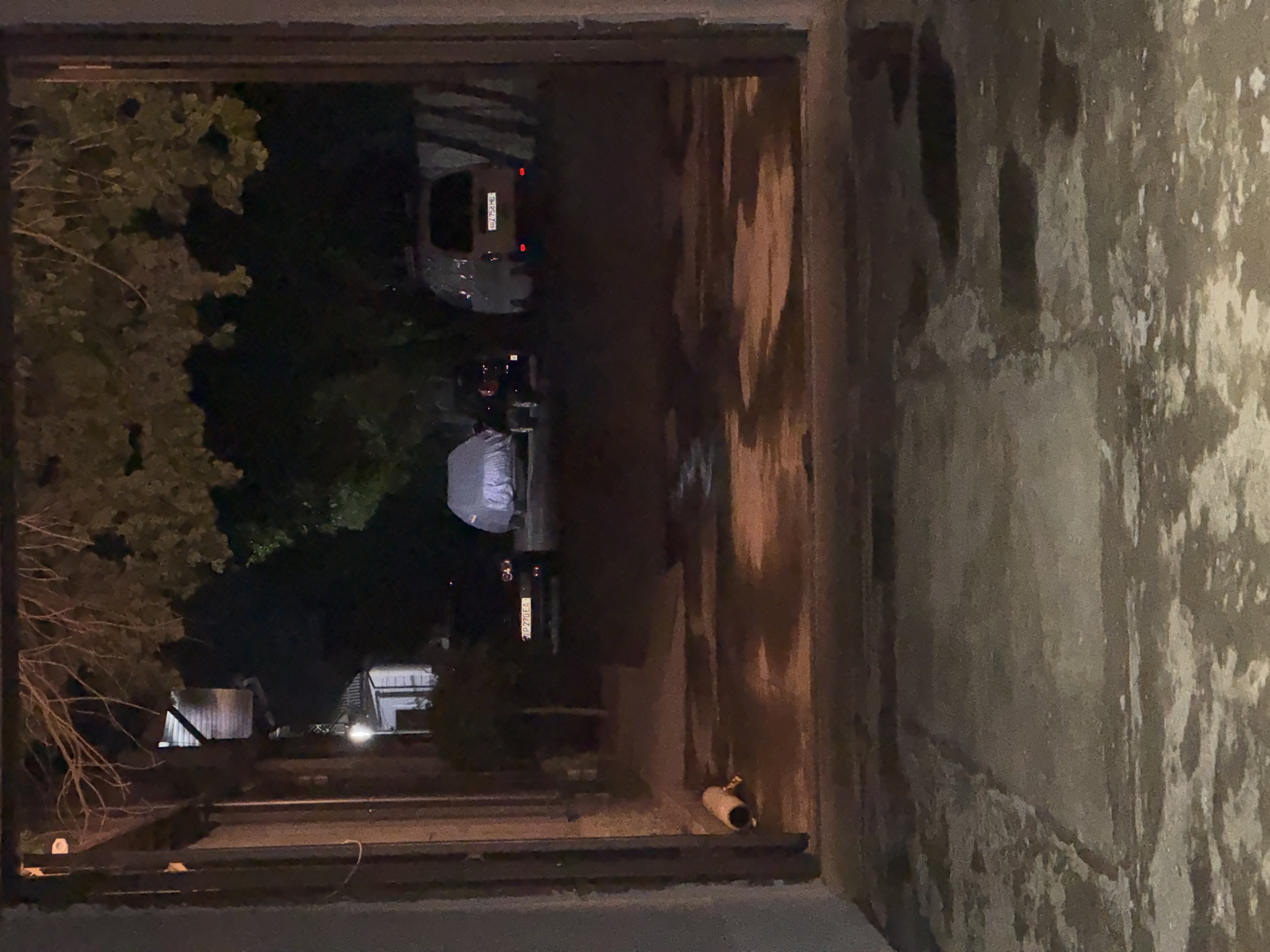 The image depicts a dimly lit outdoor scene, likely a yard or driveway, viewed through an archway. The ground in the foreground is concrete with several wet patches reflecting the ambient light. In the mid-ground, there are three vehicles. The leftmost vehicle is partially obscured and appears dark. Next to it is a car covered with a light-colored tarp. To the right, a dark-colored car with illuminated taillights is parked, displaying a license plate. Further to the right, a white van is parked, also with illuminated taillights and a visible license plate. Behind the vehicles, trees with dense foliage create a dark backdrop. The overall atmosphere is dark and appears to be nighttime. The scene is located in Yalangach, Uzbekistan.