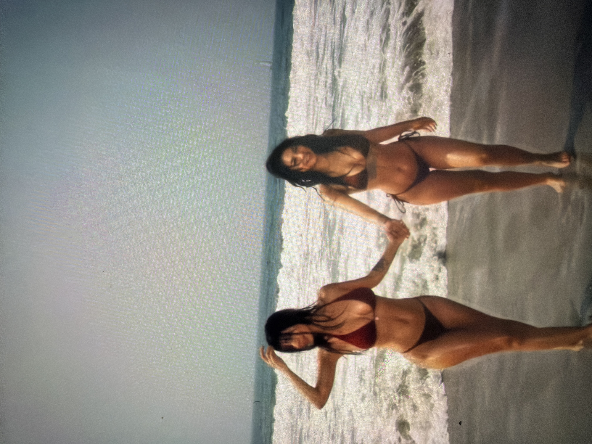 Two women are holding hands on a beach. They are both wearing bikinis. The sky is overcast. The location is not in Yerevan, Armenia, as there are no beaches in that city.