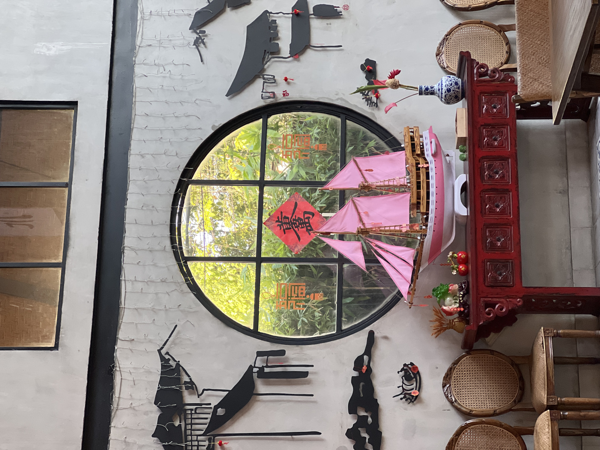 The image depicts an interior scene at a cafe in Denpasar, Indonesia. A decorative wall features black cutouts of traditional Chinese architecture, along with strings of small white lights. A large circular window offers a view of lush green foliage.
Dominating the center of the wall is a model of a pink-sailed ship. Below the ship, a bright red wooden console table displays various decorative items. These include a ceramic vase with blue and white patterns containing red and brown flowers, a small wooden box, two green decorative spheres, and a pair of red apple-shaped ornaments. To the left of the table, two ornate rattan chairs are visible. The floor in the foreground is tiled. The overall ambiance suggests a blend of cultural influences and decorative elements.