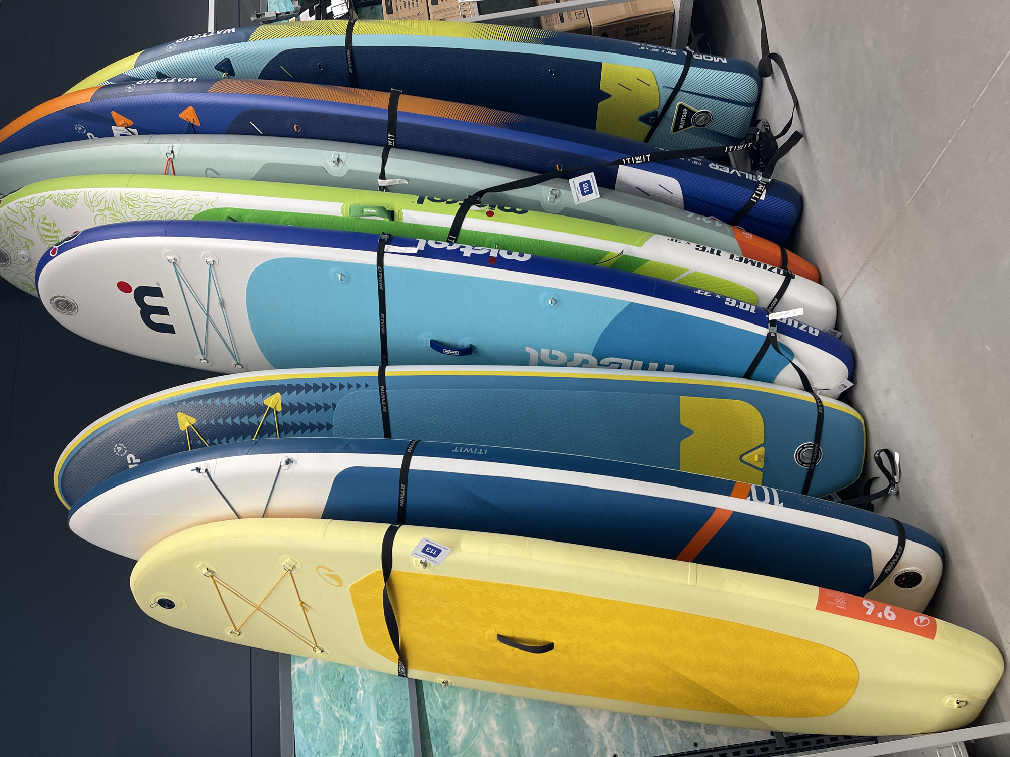 The image shows a stack of paddle boards stored indoors, likely in a retail environment. The boards are arranged horizontally on a wall rack and secured with black straps labeled "B'TWIN". The boards vary in color and design, with yellow, blue, green, and white being prominent. Some boards have printed logos and markings, including brands like "ITIWIT", "WATTSUP", "mistro", "MOR", and "3." along with size and model information such as "9.6", "100", "10'6 x 33"", "COZUMEL 11'63"", "SILVER", and "MANTA 102". A tag labeled "113" is attached to one board, and another tag displays "116". The overall setting appears to be a store or warehouse. There is no information about the location in the city of Sofia, Bulgaria in the image.