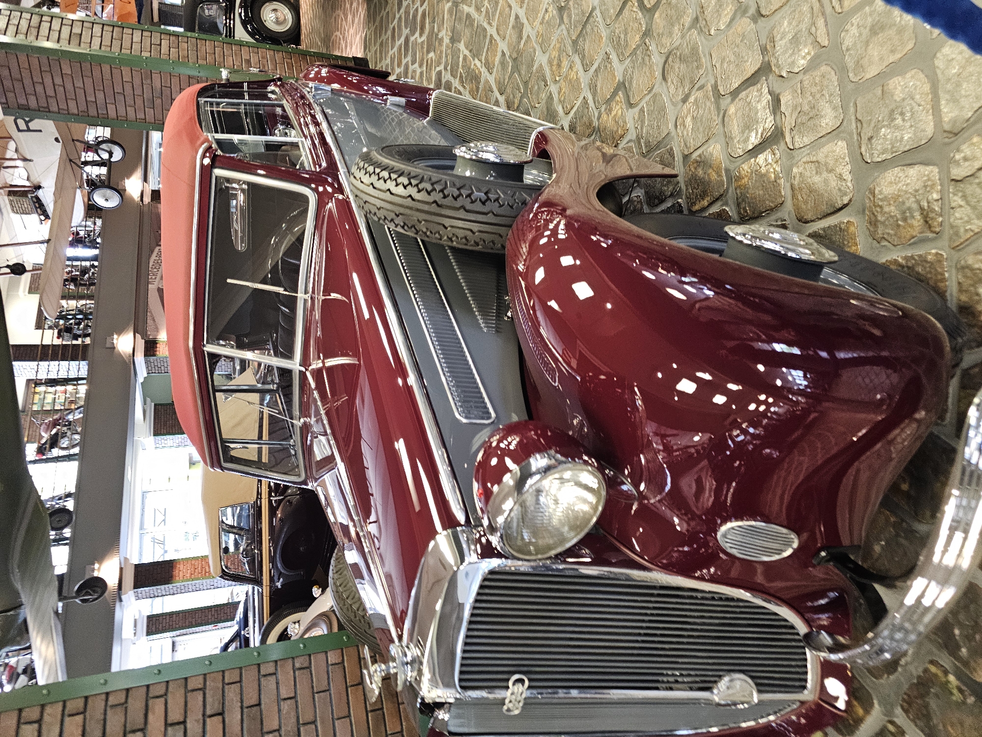 The image shows a burgundy vintage car on display at the Архангельское, Russia. The car is positioned on a stone tile floor near a brick and green-painted wall. The car features a spare tire mounted prominently on its hood and has chrome detailing on the grill and lights. The interior and windows are visible, and reflections on the car's surface suggest a polished finish. In the background, there are displays of other vehicles and historical objects visible through windows or openings.