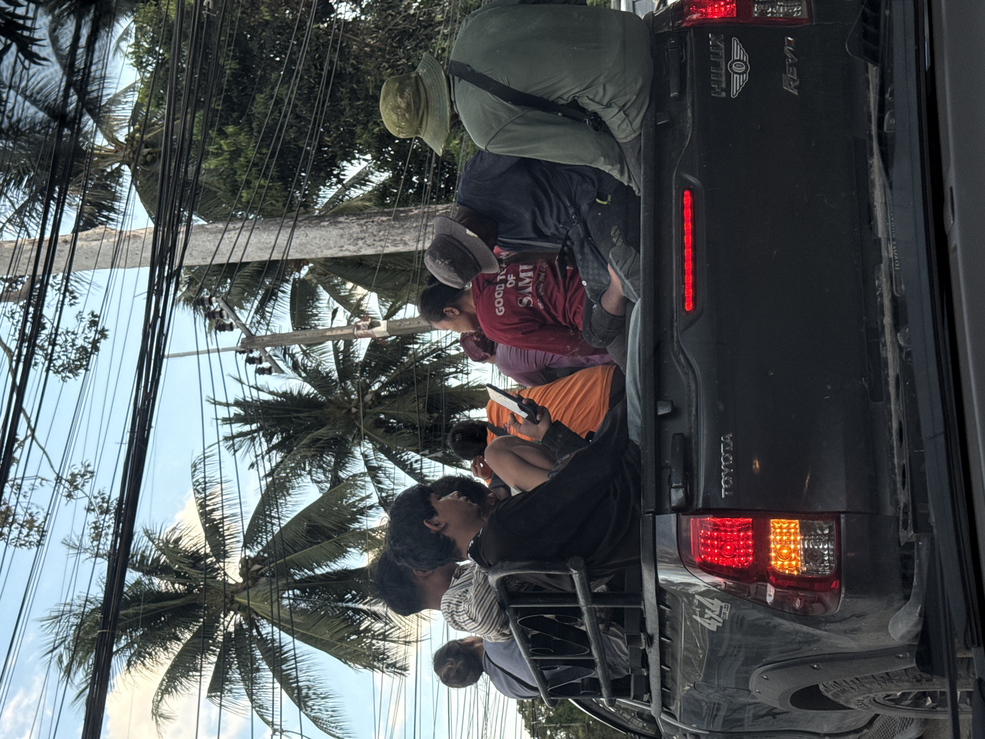 A group of people are packed into the back of a dark-colored pickup truck, likely a Toyota Hilux Revo, for transportation. The truck is in motion, indicated by the blur in the background. The scene is set outdoors, with lush green palm trees and foliage visible, suggesting a tropical or sub-tropical environment. The sky is a bright, clear blue, and the lighting suggests it is daytime, likely midday given the bright conditions. Overhead, numerous electrical wires crisscross the frame, a common sight in many parts of Thailand.
The people are diverse in age and attire. Some are wearing hats, including a wide-brimmed hat on one individual and a baseball cap on another. Clothing ranges from casual t-shirts to what appears to be workwear. The people are seated or standing in the truck bed, some looking forward, others engaged with their phones, and some simply looking around. There is a sense of shared travel and perhaps a bit of discomfort due to the crowded conditions. One person in a red shirt has text visible on it that reads "GOOD TEAM OF SAMI". Another visible text on the truck is "TOYOTA". The overall atmosphere appears to be one of daily commute or travel in a rural or semi-rural area of Thailand, specifically Baan Mae Nam. The lack of overt expressions of distress or extreme joy suggests a routine activity for these individuals.