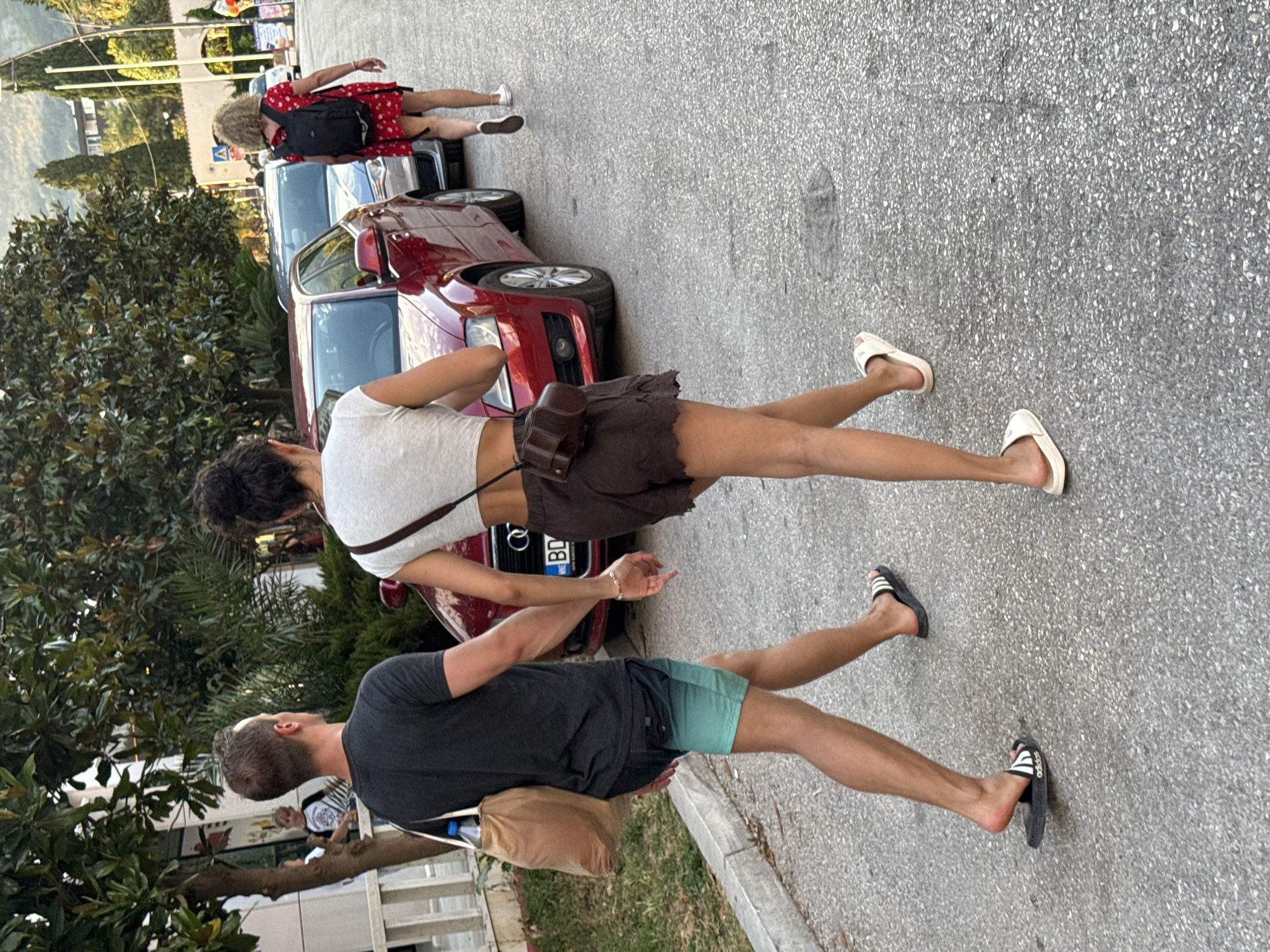 In Budva, Montenegro, a couple walks hand-in-hand down a street lined with trees and parked cars. The man, on the left, wears a dark t-shirt and turquoise and black swim trunks, carrying a beige tote bag. The woman, on the right, wears a white crop top, brown shorts, and white slides, with a brown crossbody bag. Behind them, a woman in a red polka-dot dress with a black backpack walks away from the camera. A red Audi is parked in the middle ground, with other vehicles visible further down the street. The scene depicts a casual stroll in an urban, coastal environment.