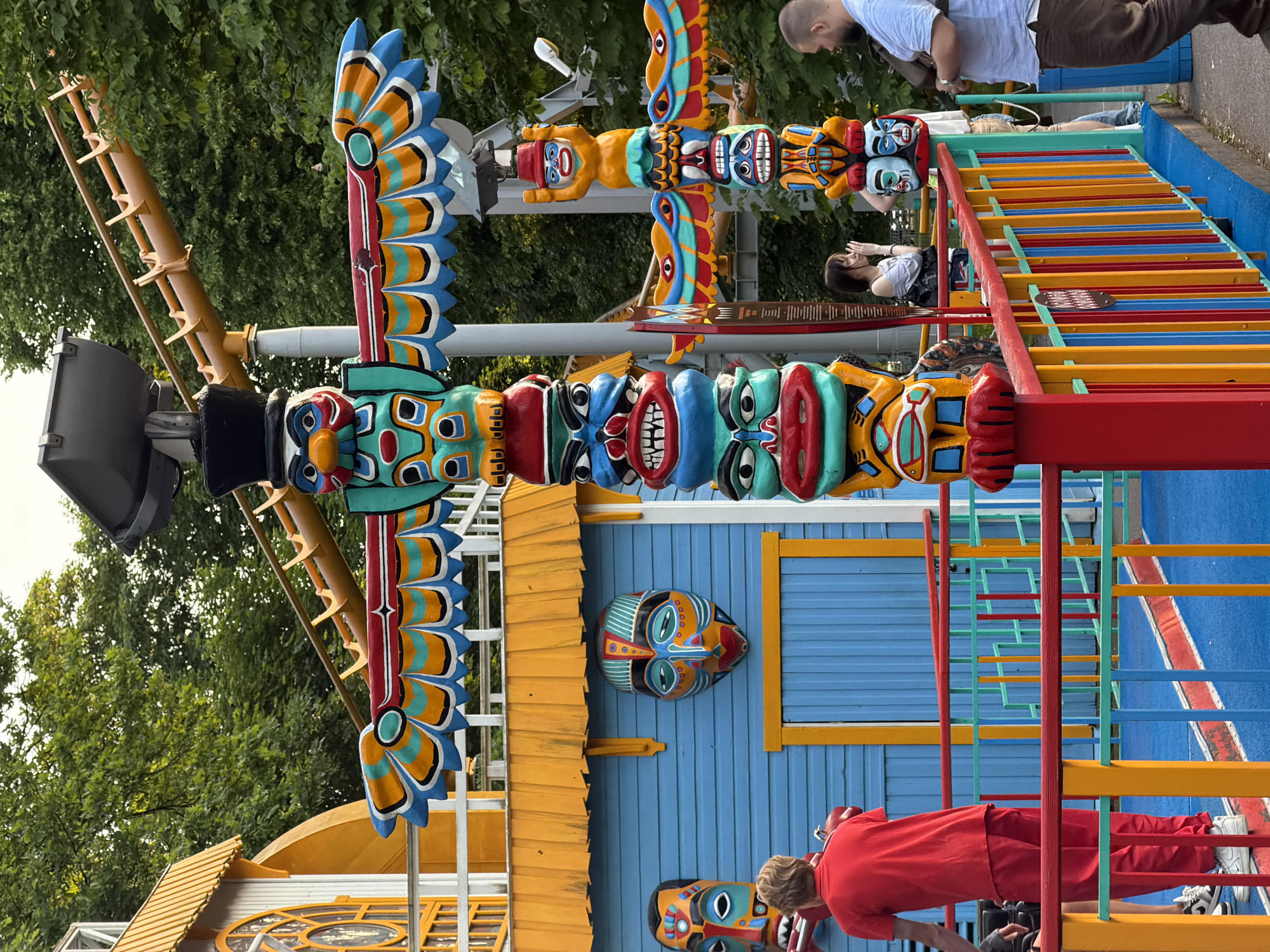 This media file shows a colorful outdoor scene, likely an amusement park or playground, with a prominent totem pole as a central object. The totem pole is elaborately decorated with vibrant colors and stylized faces or figures, exhibiting a folk art or tribal aesthetic. In the background, lush green trees suggest a warm, possibly summer day.
Several people are visible in the image, though their interactions are not entirely clear due to the composition and the orientation of the image. One person in a red outfit is captured from behind, seemingly interacting with a structure at the base of the totem pole. Other individuals, including a person in a blue shirt, are partially visible in the upper sections, suggesting a public gathering.
The setting includes a brightly painted blue building with yellow trim, a common feature in amusement or recreational areas. A colorful climbing structure, made of red, yellow, and blue bars, is also a significant element, implying an area designed for recreation. The overall atmosphere appears lively and playful, characterized by the bright colors and the presence of people. There is no visible text in the image. The specific location cues, such as the style of the totem pole and the overall environment, combined with the provided location context of Saint Petersburg, Russia, hint at a cultural or recreational attraction within the city. The time of day appears to be daytime, with adequate natural light illuminating the scene. The weather seems pleasant, suitable for outdoor activities.
