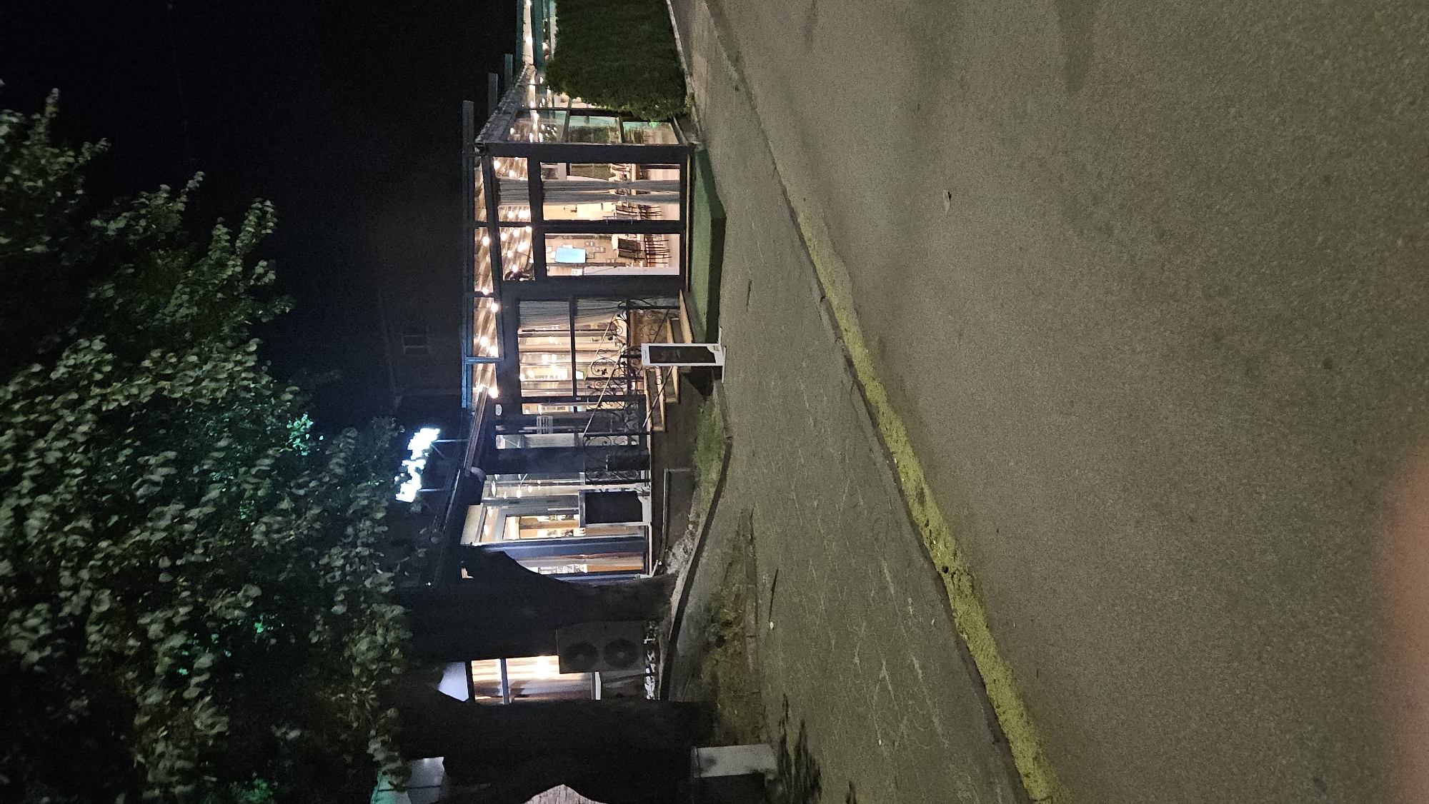 The image shows a nighttime street scene in Plovdiv, Bulgaria. A multi-story building, likely a restaurant or cafe, is prominently featured, with its interior illuminated and visible through large windows. The building is adjacent to a paved road with a yellow line marking its edge. A tree with dense foliage is located to the left of the building, partially obscuring the view. An air conditioning unit is visible at the bottom of the building. The overall lighting suggests an urban environment with streetlights or nearby illumination. No people are visible in the image.