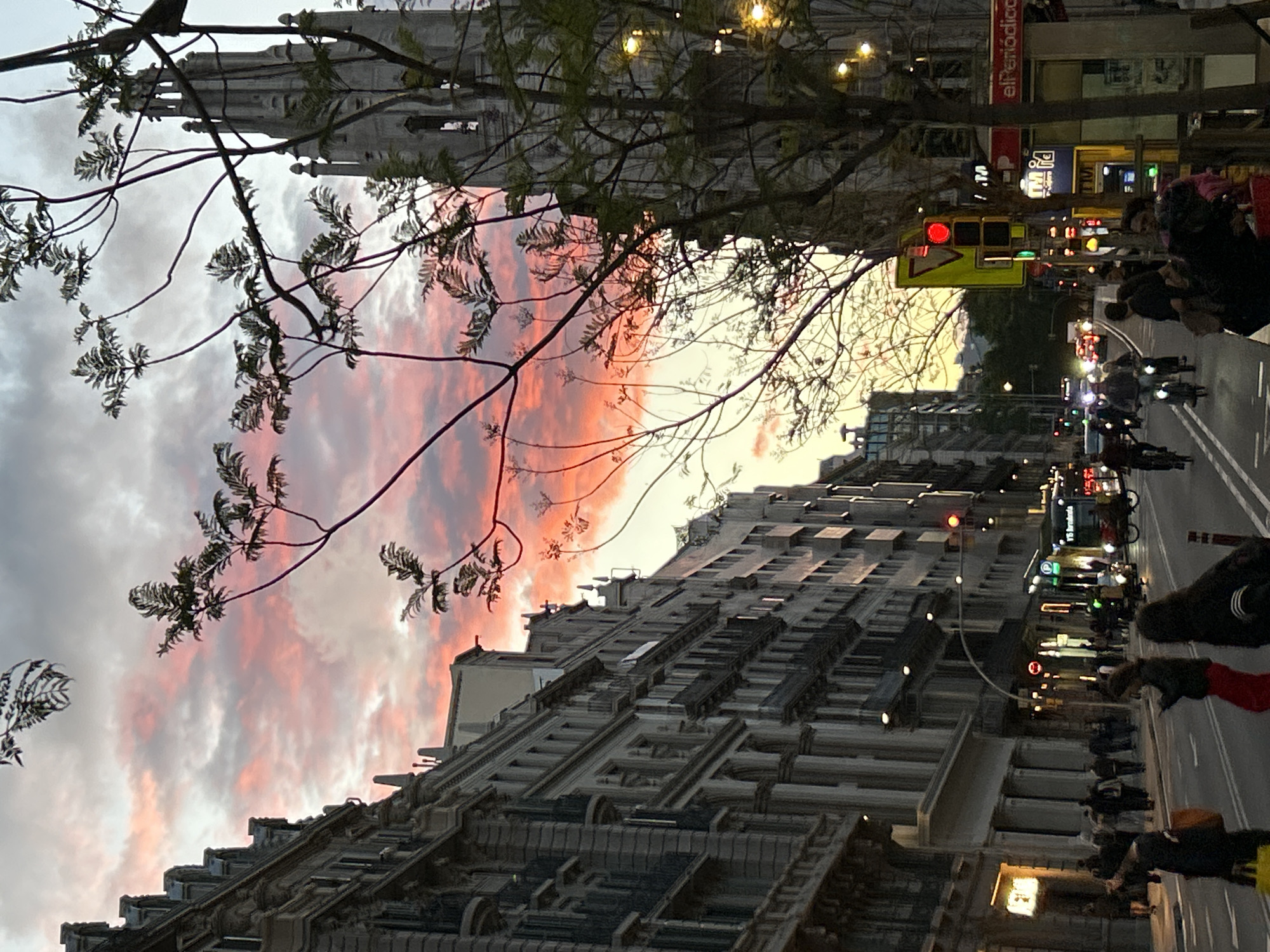 Here's a detailed description of the image:
**Scene:** An urban street scene at dusk, likely taken in Barcelona, Spain. The image is oriented at an unusual angle.
**Environment:** The sky is filled with clouds tinged with orange and pink from the setting sun. The street is lined with tall, ornate buildings, characteristic of Barcelona's architecture. Streetlights and illuminated signs cast a warm glow on the scene.
**Subjects & Objects:**
* **Buildings:** Predominantly tall, multi-story structures with detailed facades, window patterns, and cornices. A church steeple is visible in the top left corner.
* **Street:** A wide avenue with visible traffic, including scooters and possibly cars. Pedestrians are walking along the sidewalk.
* **Vegetation:** A tree with bare branches and leaves is visible in the foreground.
* **Signage:** Street signs and illuminated shop signs are visible. One prominent sign reads "El Periódico".
* **Traffic lights:** Traffic lights are visible with red lights on.
* **People:** Pedestrians are visible on the street.
**Actions & Interactions:**
* People are walking along the street.
* Traffic, including scooters, is moving along the road.
**Overall Impression:**
The image captures a vibrant urban atmosphere during sunset, with the architecture, lighting, and pedestrian activity typical of Barcelona. The presence of the "El Periódico" sign and the architectural style confirm the location.