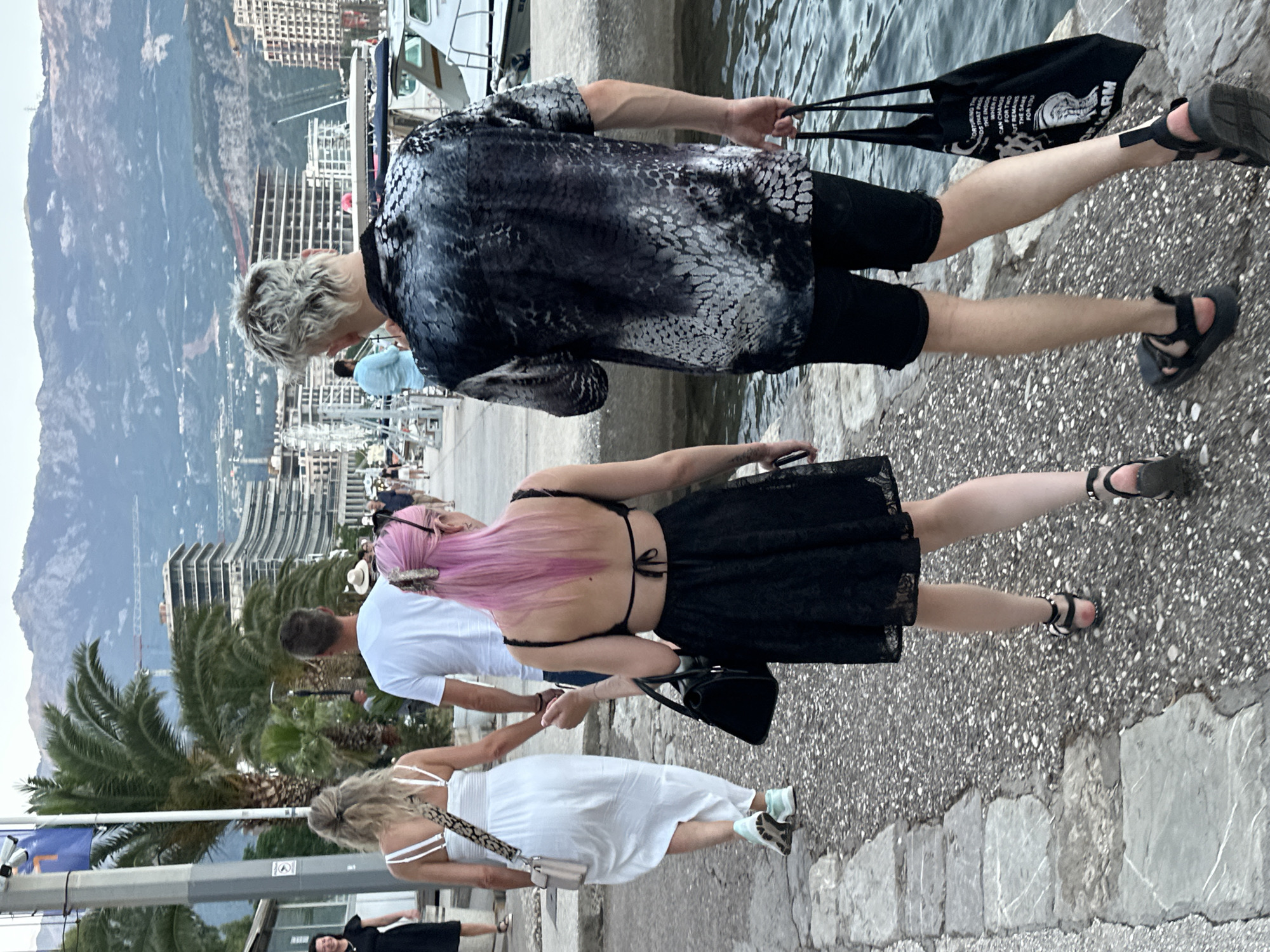 Four individuals are walking away from the camera on a paved walkway along the waterfront in Budva, Montenegro. The man in the foreground on the right is wearing a black and grey patterned shirt, black shorts, and black sandals. He is carrying a black tote bag in his right hand. To his left, a woman with bright pink hair is wearing a black lace dress and black sandals. She is carrying a black purse in her left hand. To her left, a man in a white t-shirt and jeans is holding hands with a woman in a white dress and white sneakers, who is carrying a beige purse. In the background, palm trees and multi-story buildings are visible, with mountains in the distance. The water of the Adriatic Sea is to the right of the walkway.