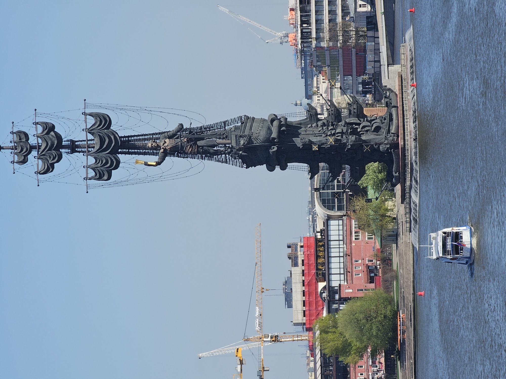 The image shows the Peter the Great statue in Moscow, Russia, situated on an artificial island in the Moskva River. The statue depicts Peter the Great standing on a ship, holding a scroll. The background includes modern buildings, construction cranes, and a passing tour boat on the river. The sky is clear and blue.