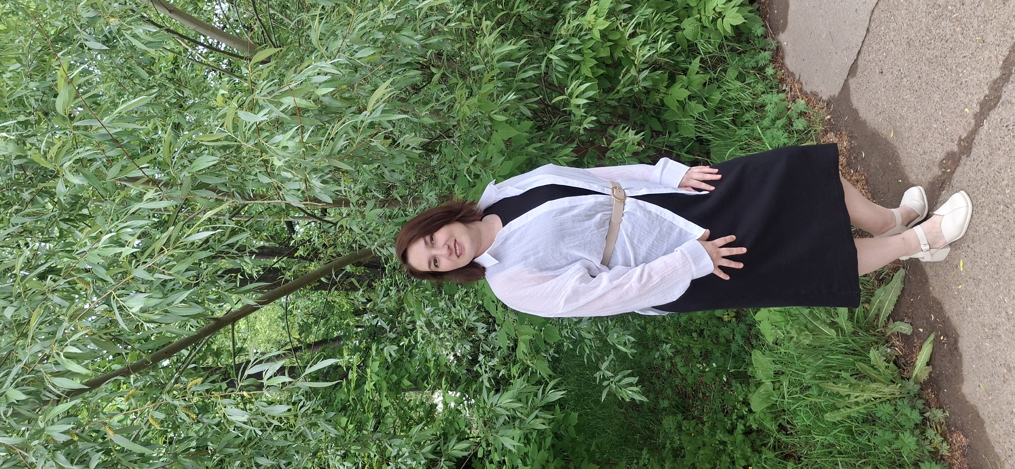 A woman stands on a concrete path next to a grassy area with trees in Chaikovsky, Russia. She wears a black dress, a white button-down shirt, a belt, and white shoes. She has brown hair and is looking at the camera. The background consists of greenery, including trees with slender leaves. The concrete path runs alongside the grassy area. The image is rotated approximately 90 degrees clockwise.