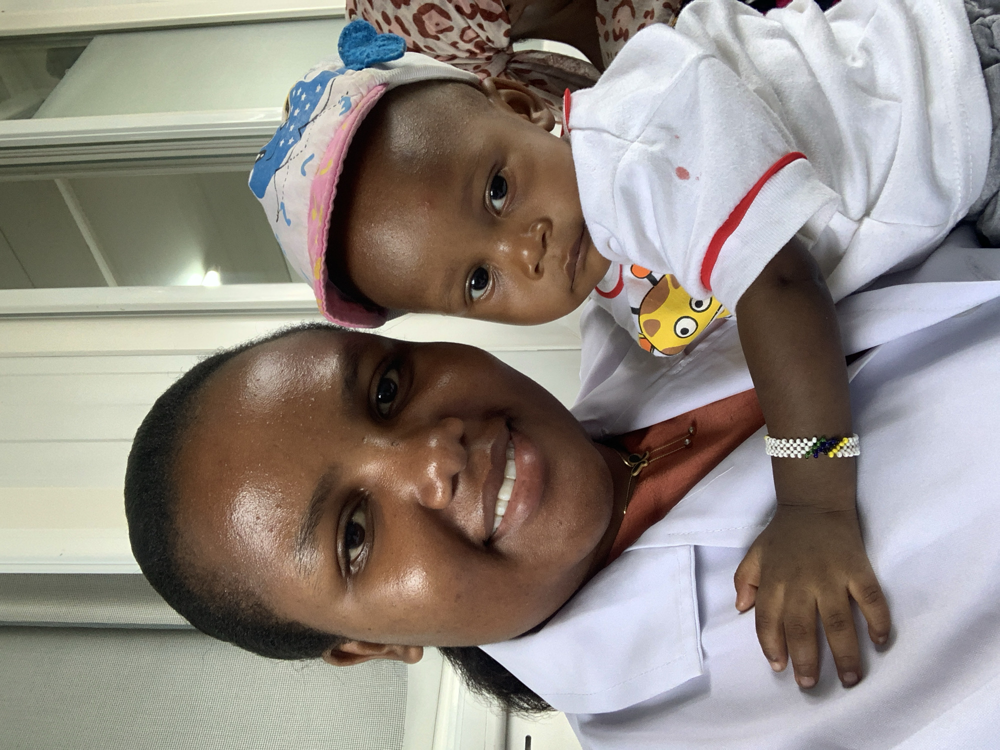 The image displays an adult female holding an infant in an indoor setting in Dar es-Salaam, Tanzania. The female, with a dark complexion and dark hair, wears a white lab coat over an orange-brown top and a gold-colored necklace with a butterfly pendant. She is smiling and looking directly at the camera. The infant, also with a dark complexion, is held on her left side, looking directly at the camera with a neutral expression. The infant wears a multi-colored cap featuring a pink brim and white crown with blue patterns, a white short-sleeved t-shirt with a yellow cartoon giraffe graphic on the chest, red trim on the sleeves, and a red spot on the right shoulder area. A white beaded bracelet with yellow, green, and blue accents is visible on the infant's right wrist, with the left hand resting on the female's lab coat. The background features light-colored window blinds or textured walls on the left and white-framed windows or glass partitions on the right, one reflecting a light source. A third individual, partially obscured and wearing patterned fabric, is visible behind the infant on the far right.