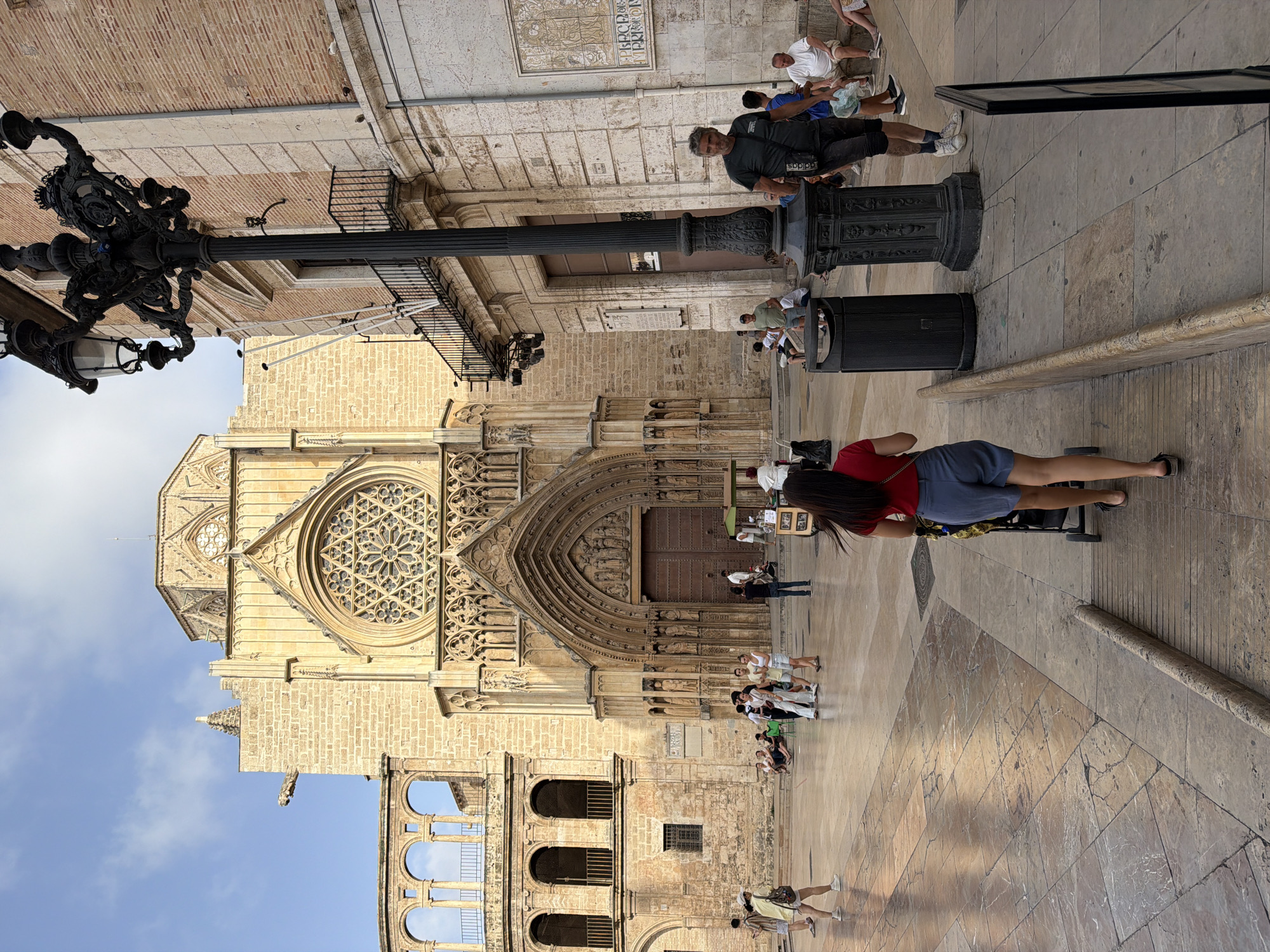 In an outdoor urban plaza, bathed in daylight under a blue sky with scattered clouds, a woman pushes a stroller away from the camera. She is positioned in the foreground, walking on a paved surface composed of large, light-colored stone tiles with some cracks. She is wearing a red shirt and blue shorts. The plaza is populated by numerous people, some sitting on steps of a large, ancient stone building that dominates the background. This building features a grand arched doorway, a circular rose window, and ornate architectural details. To the right of the building, a tall, black, decorative lamppost stands prominently. Several trash receptacles are visible near the lamppost. In the mid-ground, other individuals are walking and standing, including a man in a dark t-shirt and shorts walking towards the right, and a group of people sitting on the steps of the building. Further to the left, a woman in a yellow top and shorts walks away from the camera. The overall scene depicts a bustling public space with a significant historical architectural element.