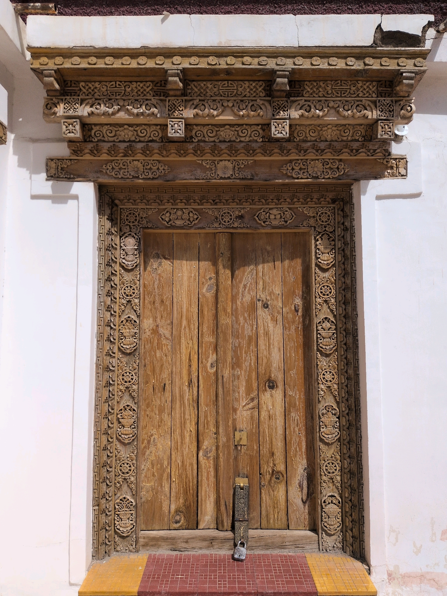 The image shows a closed, weathered wooden double door with intricate carvings around its frame and a decorative wooden frieze above it. The door is secured with a metal lock and has a tarnished brass latch. The wall surrounding the door is painted white, with some areas showing signs of age and repair. Below the door, there is a step with red and yellow tiling. The architectural style suggests a traditional building in a region with rich cultural heritage. The scene is well-lit by natural sunlight, casting shadows that highlight the detailed carvings. The image is taken from a frontal perspective, at eye level, focusing on the entrance. This particular doorway is located at the entrance to the Martselang Monastery, Martselang, India.
