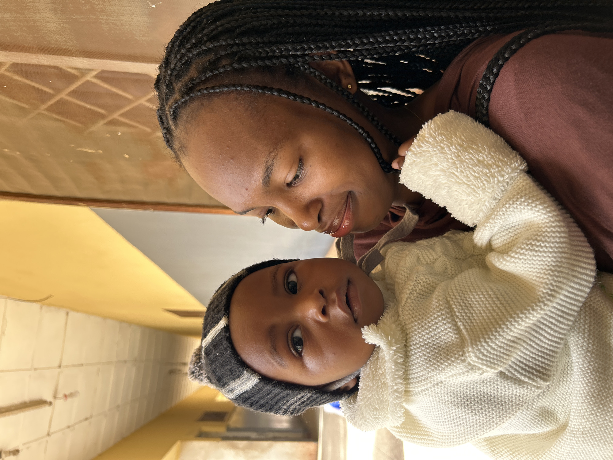The image shows a young child being held by an older individual. The child is wearing a striped knit hat and a cream-colored sweater. The older individual has braided hair and is looking down at the child. They are standing in what appears to be a hallway with yellow walls and a dropped ceiling with fluorescent lighting. A patterned grate is visible on the wall in the background. The scene is likely in Jos, Nigeria, given the context of the prompt, but there are no explicit visual cues within the image to confirm this specific location.