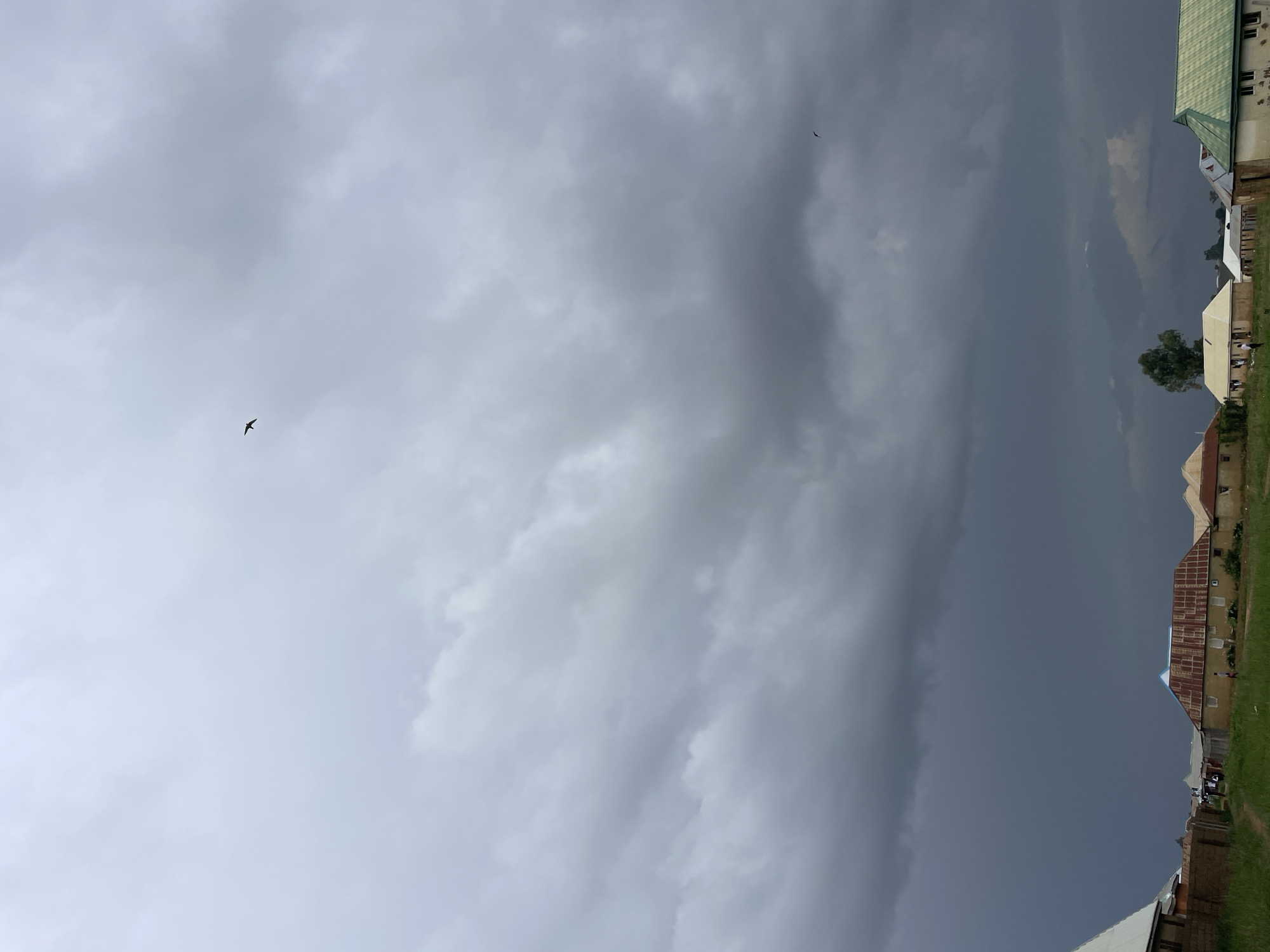 The image presents a partially cloudy sky, with a clear delineation between the bright, uniform light of the clouds to the left, and an approaching dark storm front to the right. Two birds are visible. The buildings and landscape of Durimi, Nigeria are visible on the right edge of the frame.