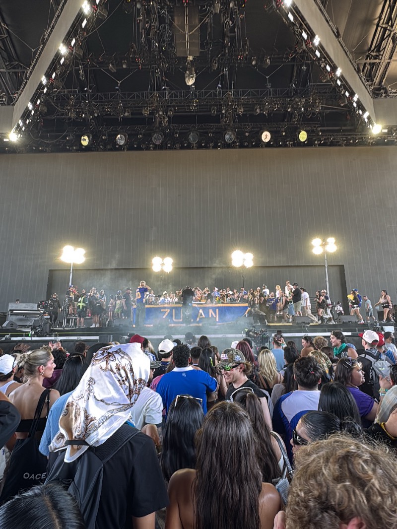 The image shows a crowd of people at a concert in Palm Desert, United States. The crowd is facing a stage where a performance is taking place. Stage lights illuminate the performers, whose appearance suggests a rap concert. A large, plain backdrop is situated behind the stage. Overhead rigging and lighting equipment hang from the venue's ceiling.
