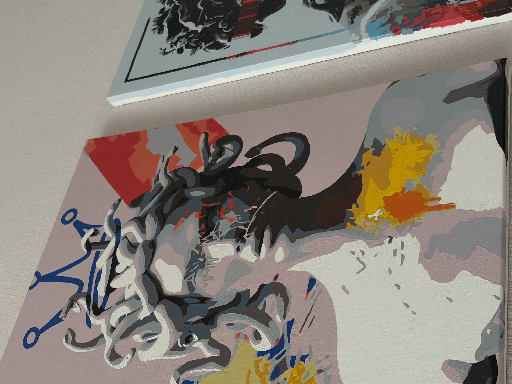 Two paintings are hung on a wall. The first painting, visible in the upper portion of the frame, has a light-colored background and a dark, complex design. Below it, another painting features a pink background with an abstract combination of colors and shapes, with prominent elements of blue, white, yellow, and gray.