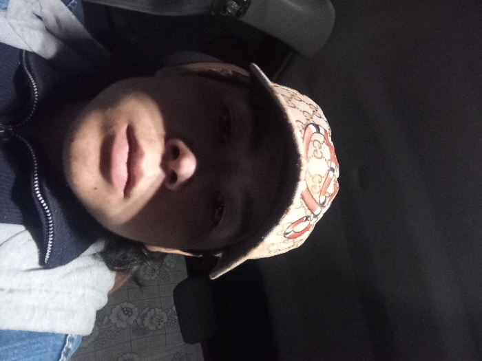 The image depicts a male individual, appearing to be a young adult, in a dimly lit, enclosed indoor setting within the city of Konkus, Uzbekistan. The subject is captured from the chest up, looking directly towards the camera.
He wears a beige baseball cap adorned with a repeating pattern of red and black intertwined serpentine motifs against a tan background of stylized 'C' or oval shapes. His dark hair is visible on the right side of his neck. He is dressed in a dark blue quarter-zip collared garment with a silver zipper, beneath which a light grey hooded garment is partially visible on his shoulders and chest. A portion of a blue denim-like jacket or shirt is also visible on his right shoulder.
Lighting emanates from the upper right (viewer's perspective), casting the subject's forehead and eyes into shadow while illuminating his nose, mouth, and chin. The background consists of a uniform dark grey or black surface above, likely a ceiling. To the left, a light grey or off-white vertical element is present. To the right, behind the subject's shoulder, a patterned surface features large white floral designs, resembling roses, against a darker, possibly blue-grey, background. No specific actions or interactions beyond posing for the photograph are evident.