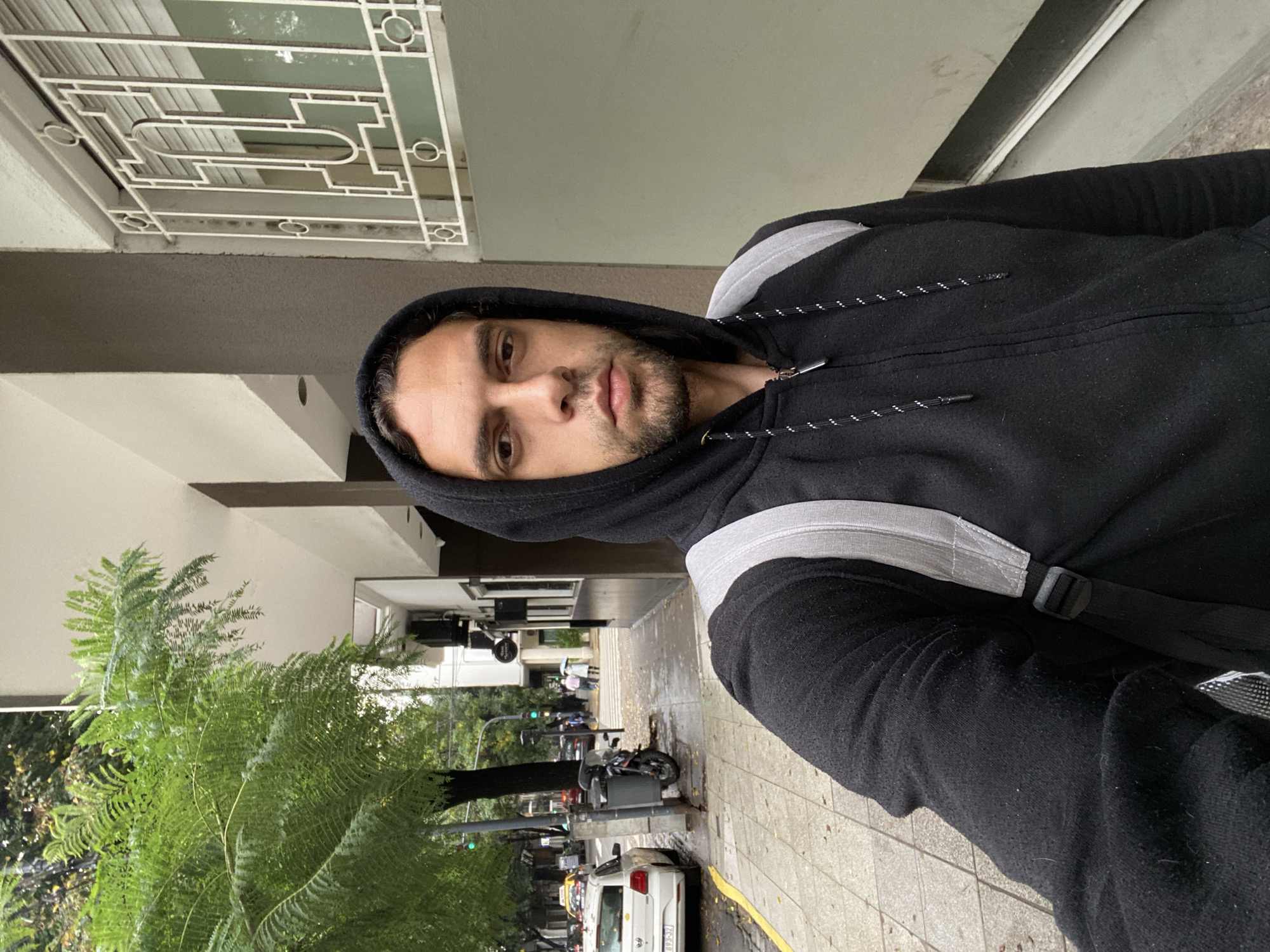 A casual portrait captures a man taking a selfie outdoors in Buenos Aires, Argentina. The man, with dark hair and a light beard, looks directly at the camera with a neutral expression. He is wearing a black hooded sweatshirt with light gray stripes on the shoulders and a black backpack strap visible over his right shoulder.
The image is presented in a landscape format, rotated 90 degrees clockwise from its original portrait orientation. The background reveals an urban street scene on what appears to be an overcast day, possibly after rain, as the ground in some areas looks wet and people in the distance are seen holding umbrellas.
To the upper left of the man (originally above him), part of a building is visible, featuring a window with a decorative white metal security grate. To the right of the man (originally below him), a city street and sidewalk stretch into the distance. The sidewalk is paved with gray tiles, and vibrant green fern-like trees line the street. A white car with an Argentinian license plate ("AA 193-SZ") is parked along the curb, alongside what might be a taxi and a motorcycle. In the mid-ground, other pedestrians are visible, and buildings line both sides of the street. A traffic light can also be seen. The overall mood is mundane, capturing a moment from daily city life.