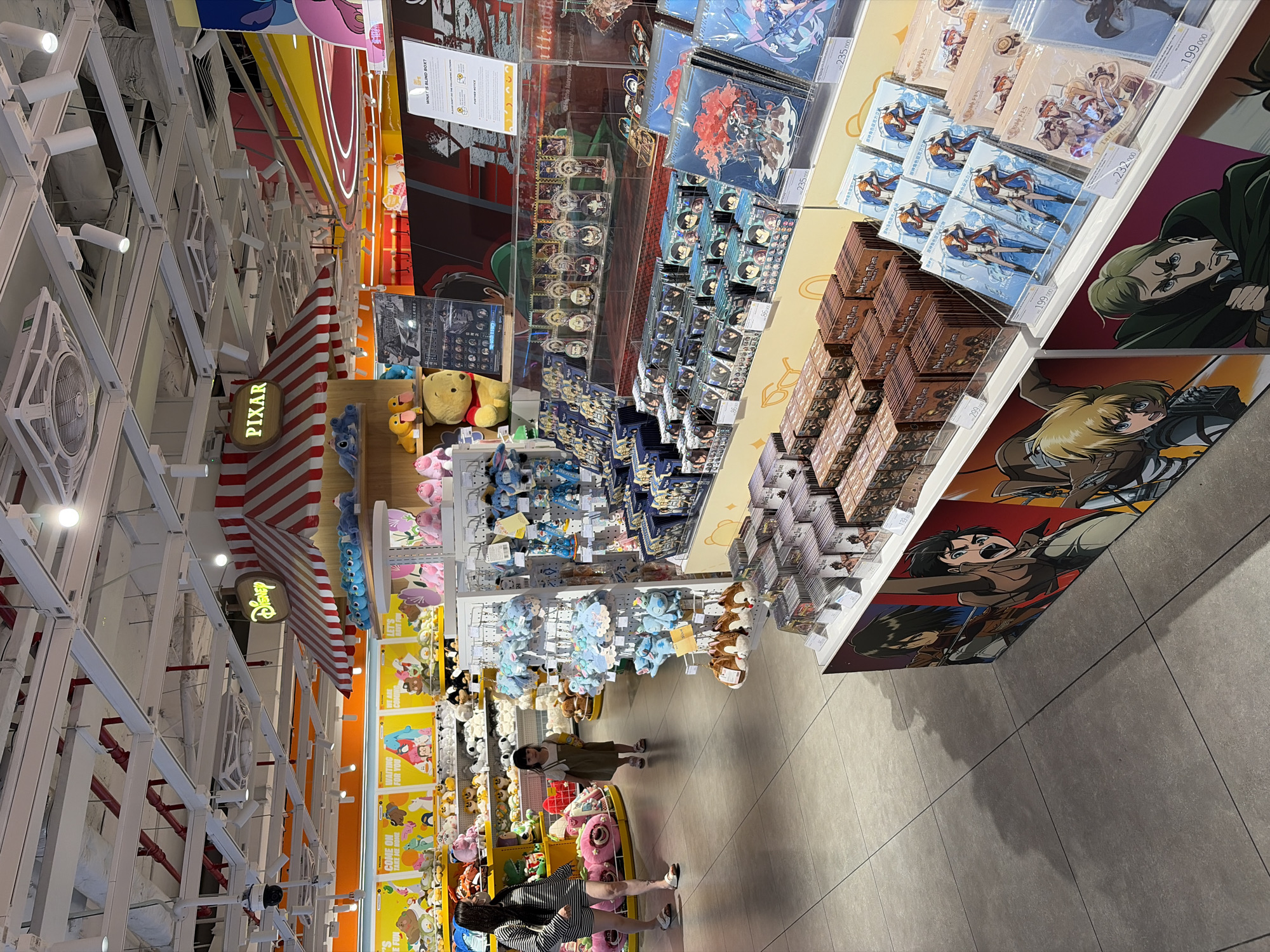 This is an indoor shot of a retail store likely located in Thủ Đức, Vietnam (according to some OCR info). The store is well-lit with a high ceiling that has white metal beams and structures. There is red-and-white striped decoration hanging from the ceiling, along with signs like "Disney" and "PIXAR."
The store's inventory is diverse, including plush toys of Disney characters like Winnie the Pooh and Stitch. In another section, there are shelves stocked with merchandise from anime series like "Attack on Titan," with character posters and merchandise bearing images of characters such as Armin Arlert and Levi Ackerman. Additionally, there are products featuring characters from "Genshin Impact" on display.
Several people are visible in the store. At least three individuals are captured in the lower part of the frame, including a woman pushing a shopping cart. One woman is standing, looking at the merchandise.