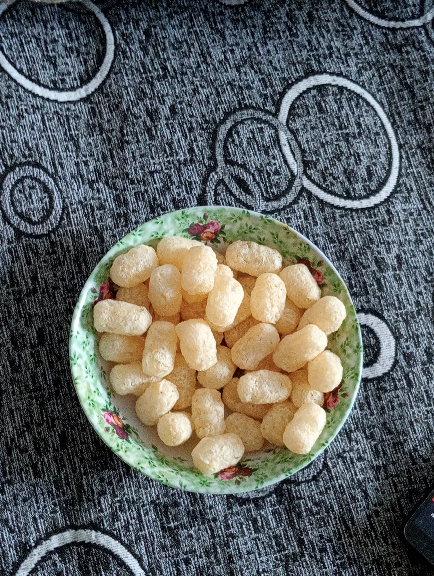 A top-down, eye-level shot shows a bowl filled with light-colored, puffed snack foods. The bowl is green with a floral pattern around its rim. The snacks are oval-shaped and appear to be made of a light, airy dough. The bowl is placed on a dark gray patterned surface, possibly a tablecloth or placemat, with a repeating circular pattern in a lighter gray. The lighting suggests it is daytime, and the setting appears to be indoors. There are no people visible in the image. The snack foods are the central focus of the image.